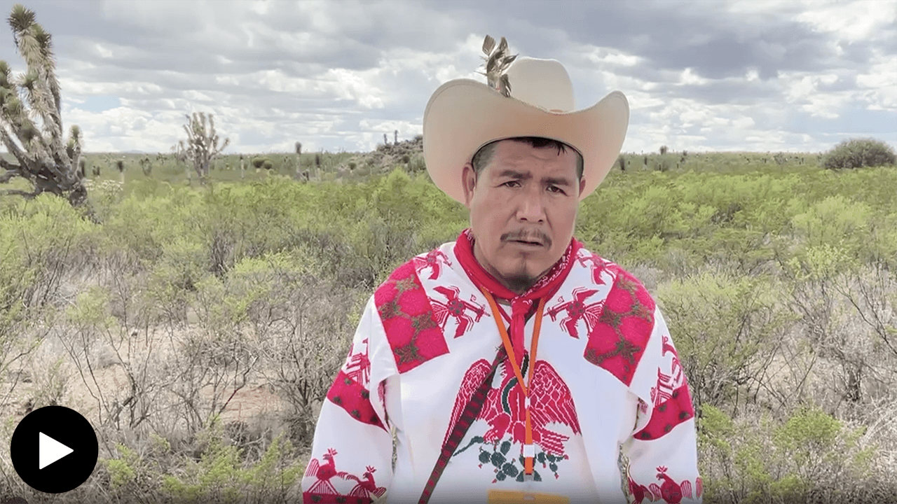 Mario Muñoz Cayetano standing in the middle of a grassy field. He looks past the camera, wearing a woven white shirt with red embellishments and designs around the shoulders and on his chest. A red bandana and lanyard are wrapped around his neck, while a curved-brim hat sits atop his head.