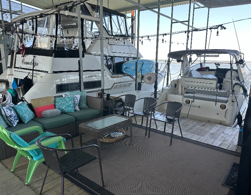 A cozy dockside seating area features wicker furniture with colorful cushions, surrounded by two white yachts moored under a covered canopy, creating a relaxing waterfront atmosphere.