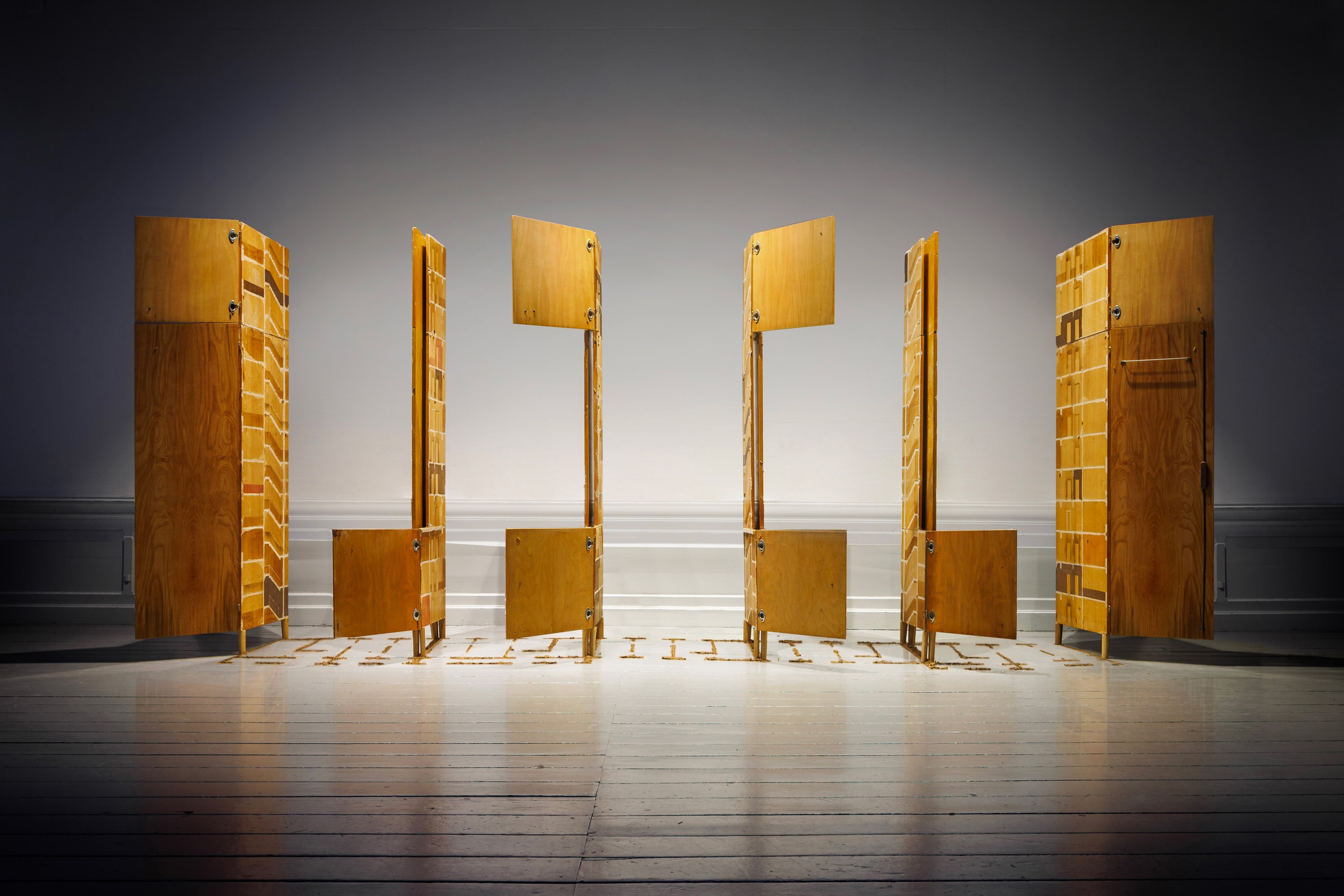 Row of tall wooden cabinet-like structures standing in a gallery space with doors partially open; sawdust is arranged on the floor beneath them in linear patterns.