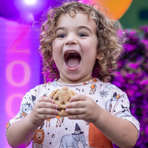 Child with curly hair excitedly holding a cookie while wearing an animal-print shirt, with colorful balloons in the background.