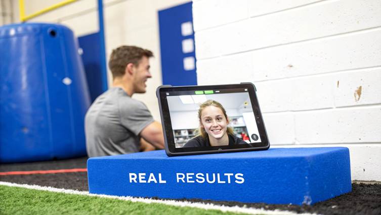 Tablet displaying a smiling woman on a video call, placed on a blue block with 'REAL RESULTS' in a gym.