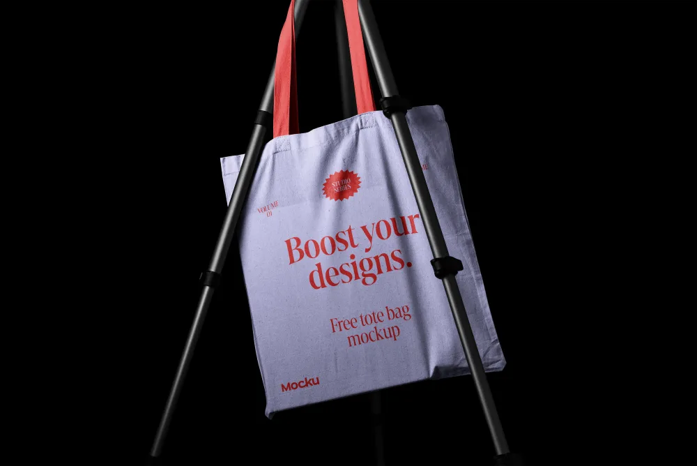 Tote bag mockup hanging on a tripod with a lavender fabric and red handles. The design features bold red typography, set against a black background for contrast.
