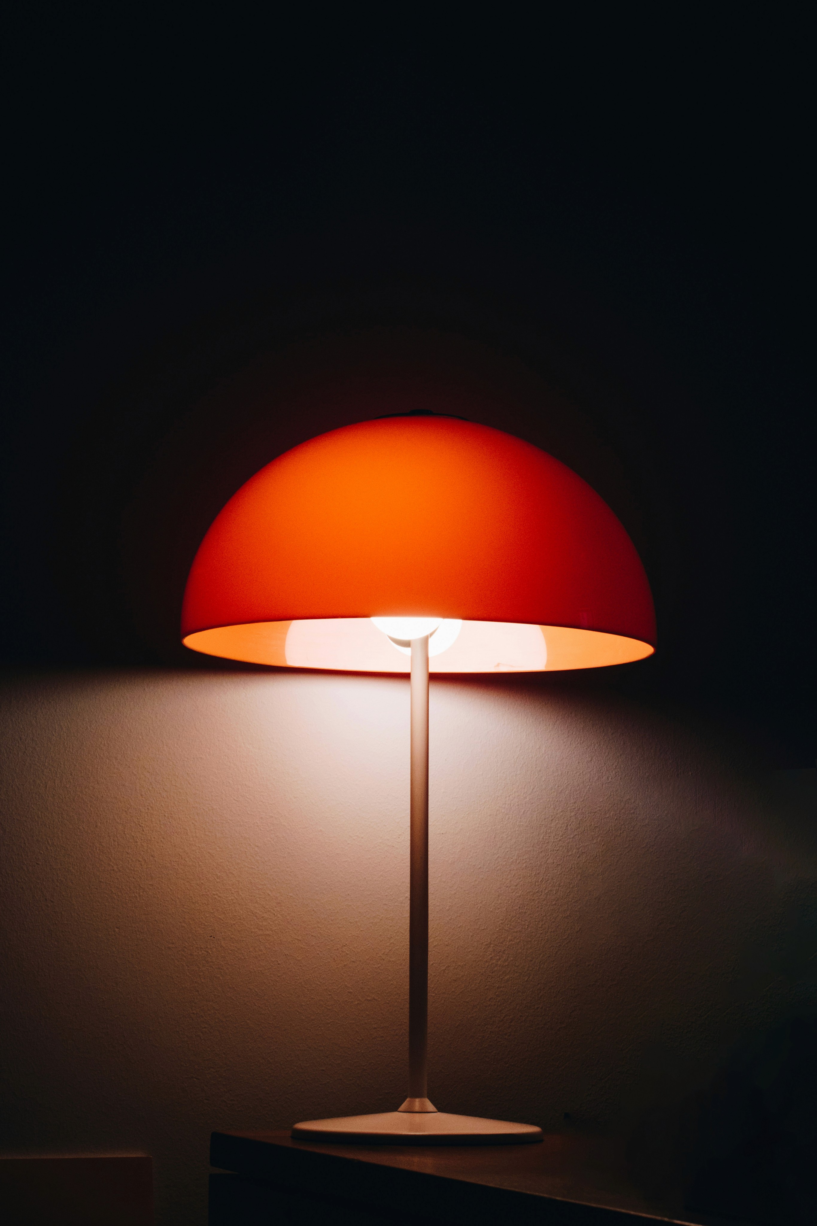 Eine orangefarbene Lampe mit einem runden Lampenschirm steht auf einem dünnen Ständer vor einem dunklen Hintergrund. Das Licht der Lampe beleuchtet den unteren Teil des Lampenschirms und den Bereich darunter.