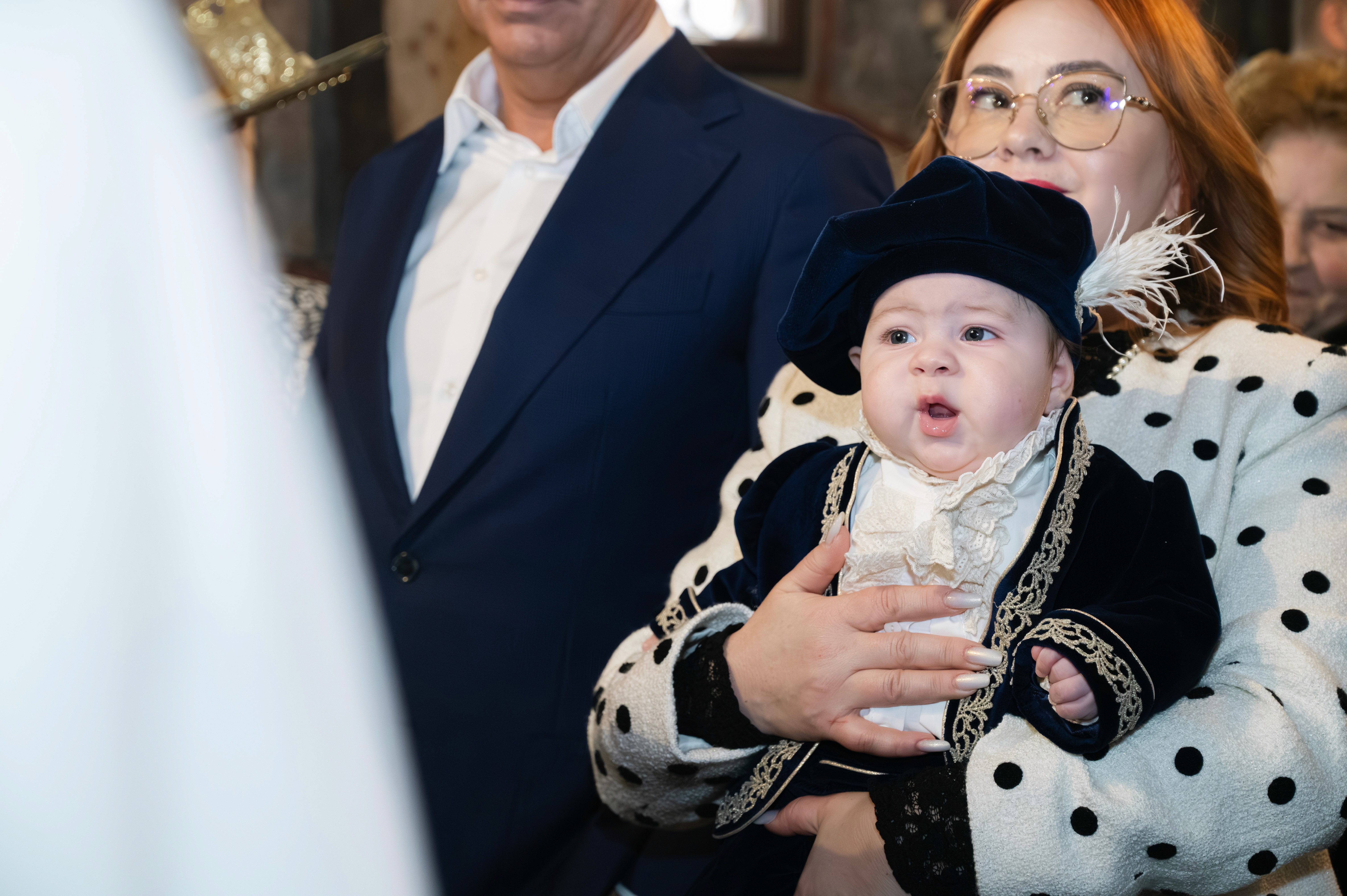 Bebeluș îmbrăcat tradițional după botez, ținut în brațe de familie.