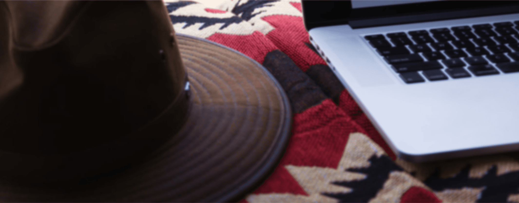 Close-up of a brown wide-brimmed hat and a laptop on a red patterned blanket.