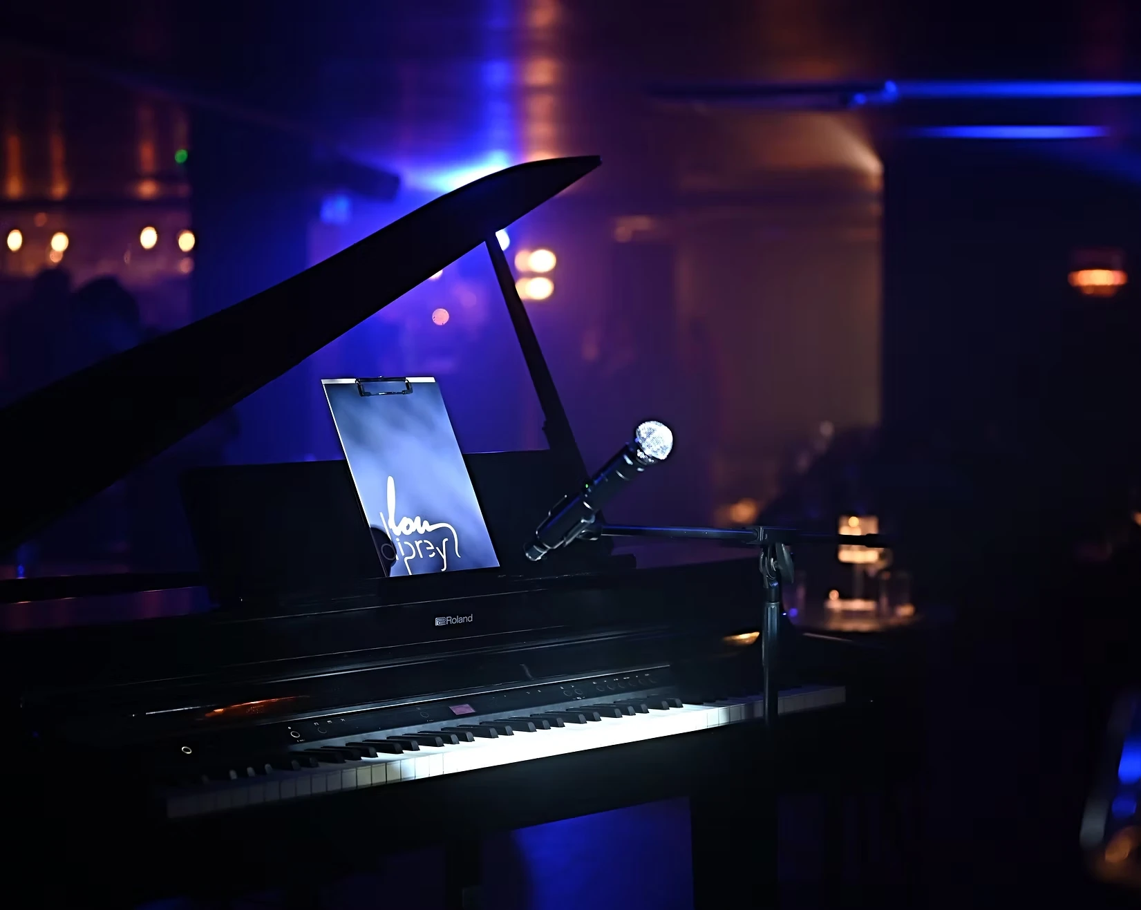 A black grand piano on a dimly lit stage with a microphone stand and a lit sign displaying the Lou Diprey logo.