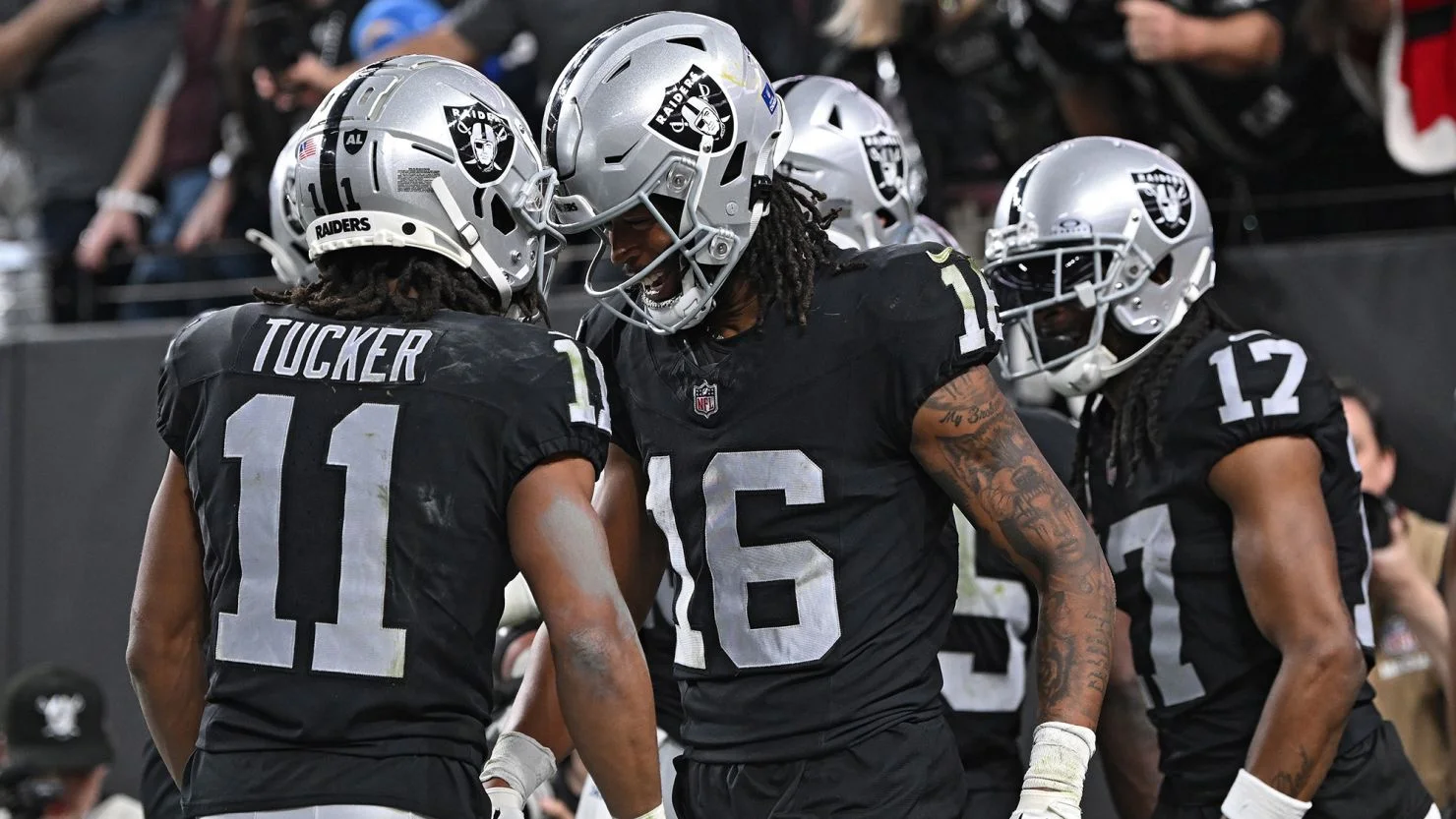 Las Vegas Raiders players in black uniforms huddle and celebrate on the field. Player #16 faces #11 “Tucker” as they lean in with intensity, while another teammate stands behind them in a silver helmet during a game.