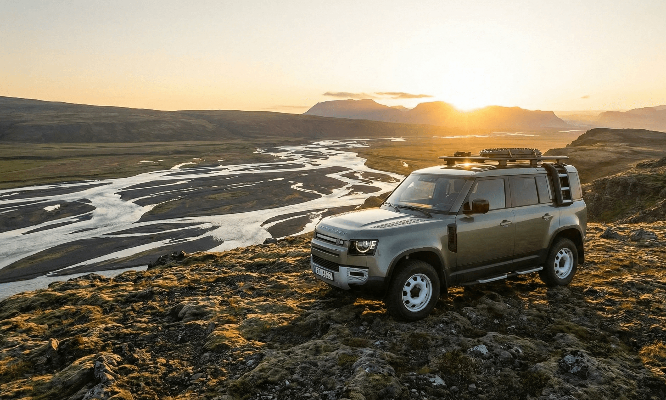 A light green Land Rover Defender with a roof rack parked on a mossy hill overlooking a winding river valley at sunset.