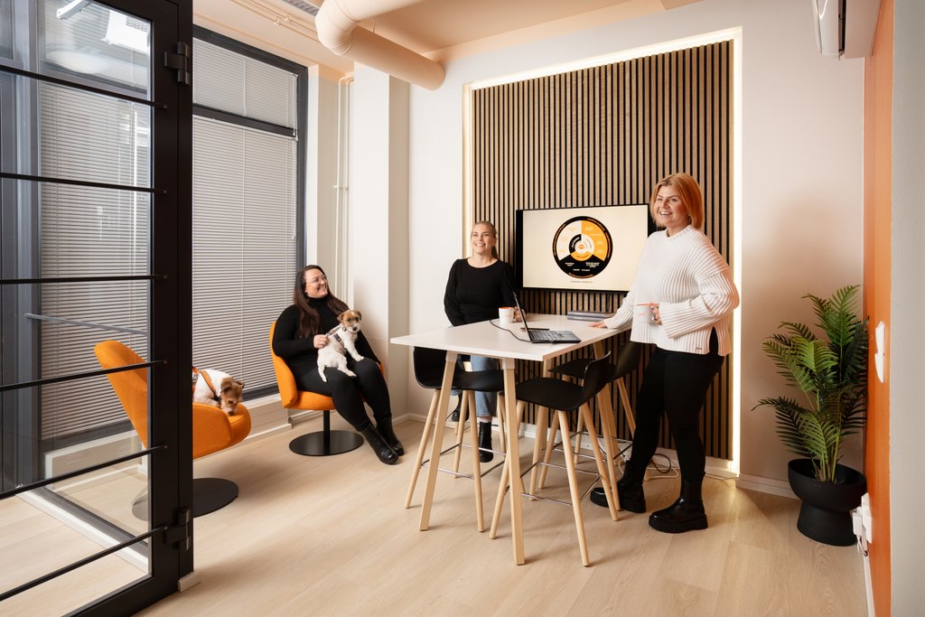 Kolme asiantuntijaa mustissa asuissa valkoisen pöydän ääressä; läppärit, mukit ja muistiot – taustalla TV:ssä social selling -kaavio; moderni ja ammattimainen valmennusympäristö.