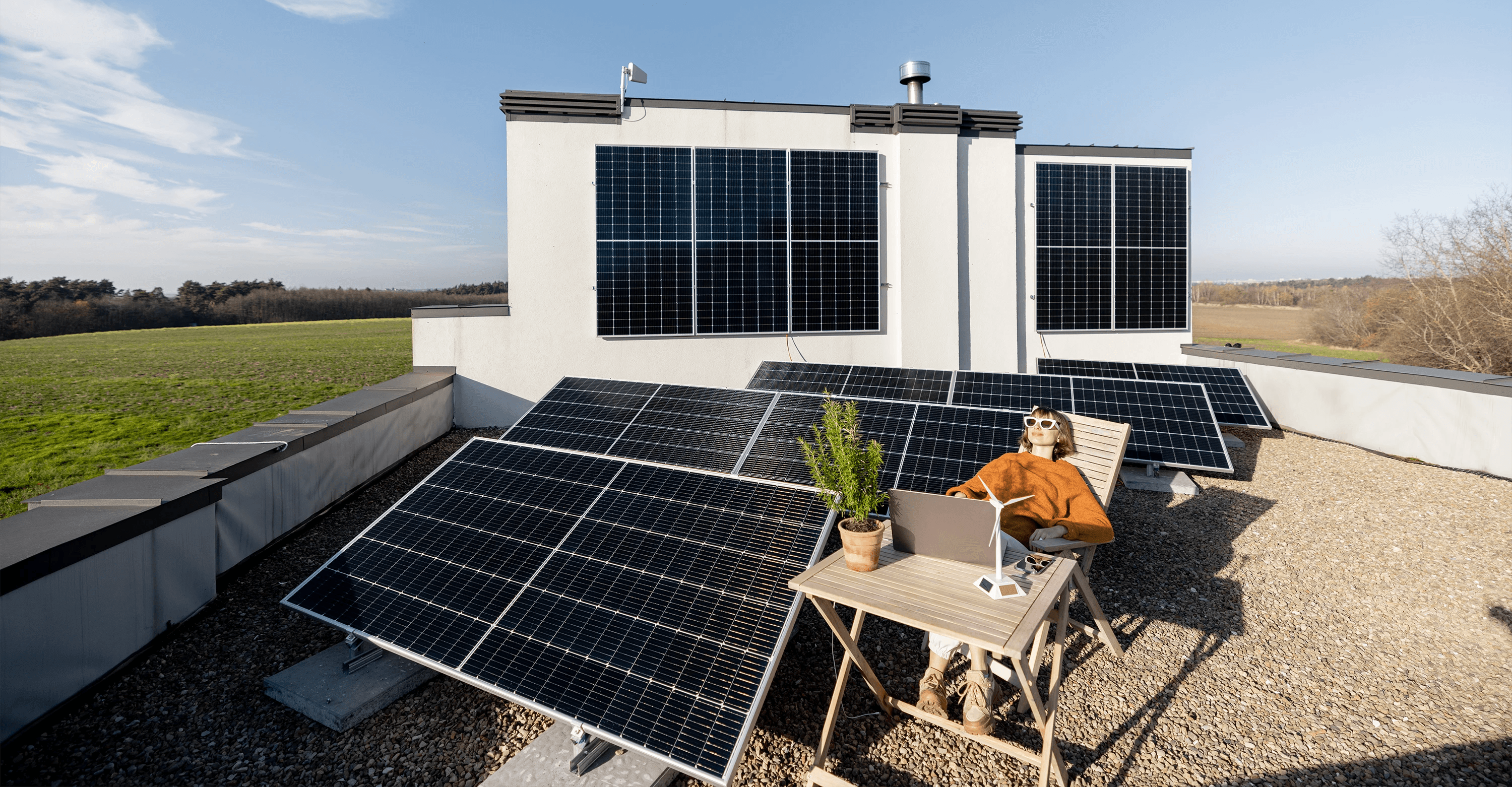 Eine Person entspannt sich auf einem Dach, umgeben von zahlreichen Sonnenkollektoren, arbeitet an einem Laptop, während im Hintergrund unter einem strahlend blauen Himmel eine weite grüne Wiese zu sehen ist.
