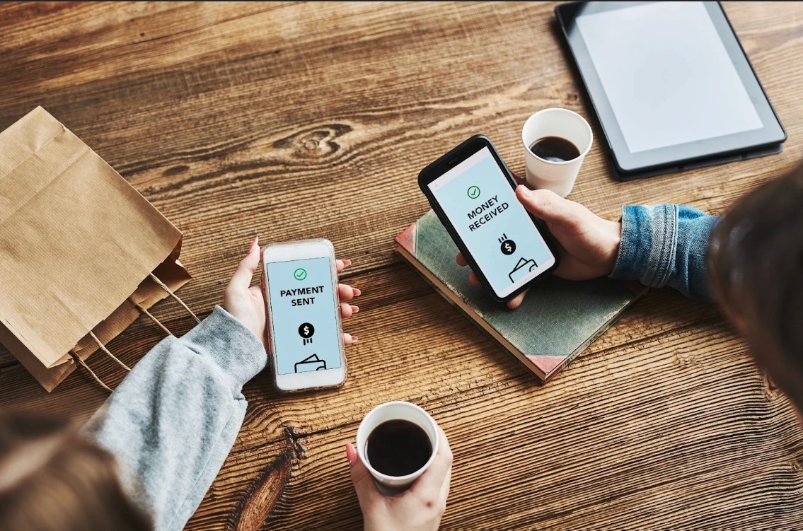  Two women transferring money with their phones and holding cups of coffee. 