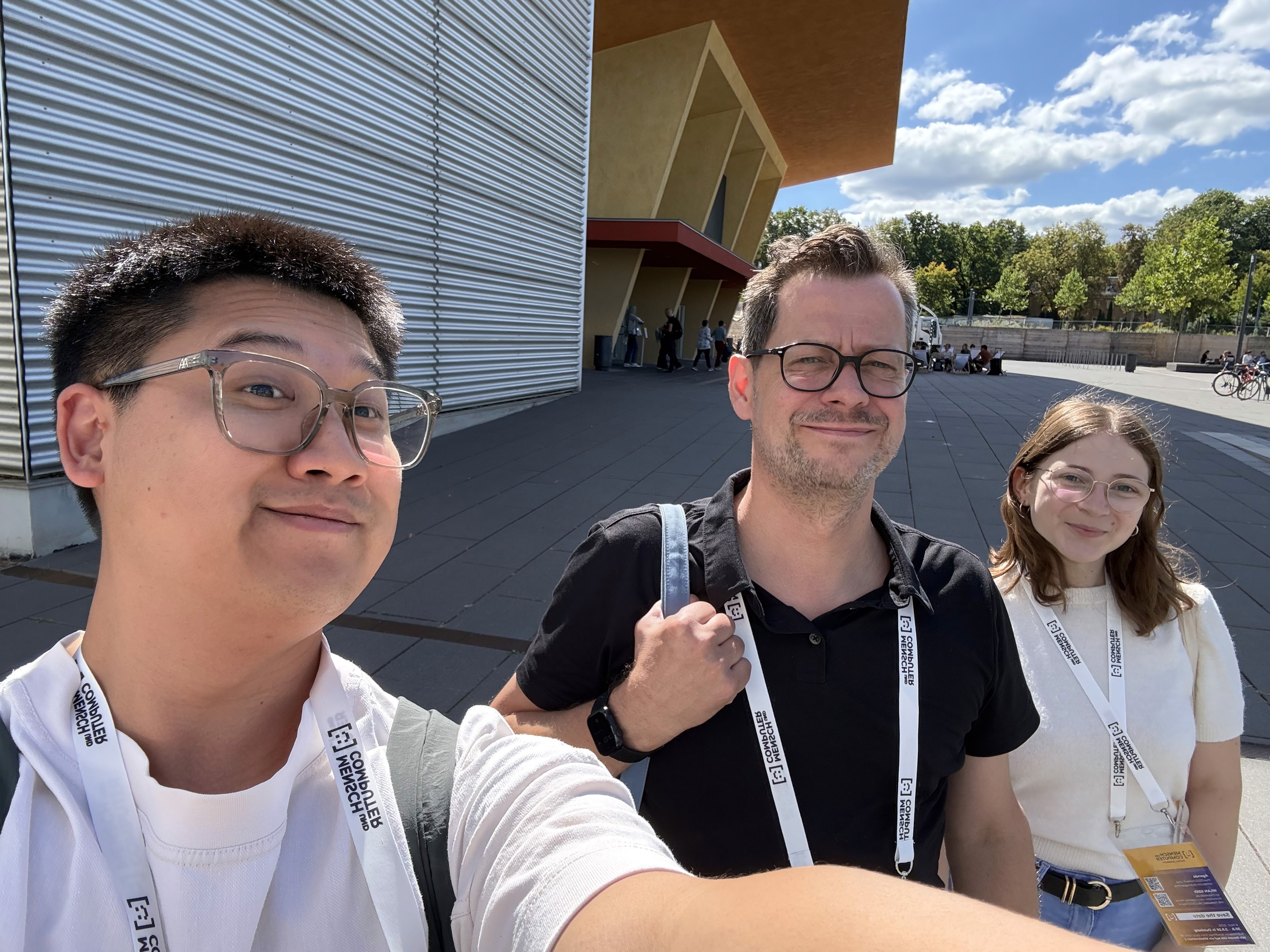 Drei Personen mit Konferenz-Ausweisen vor einem modernen Gebäude mit Metallfassade und orangefarbenem Dach bei sonnigem Himmel