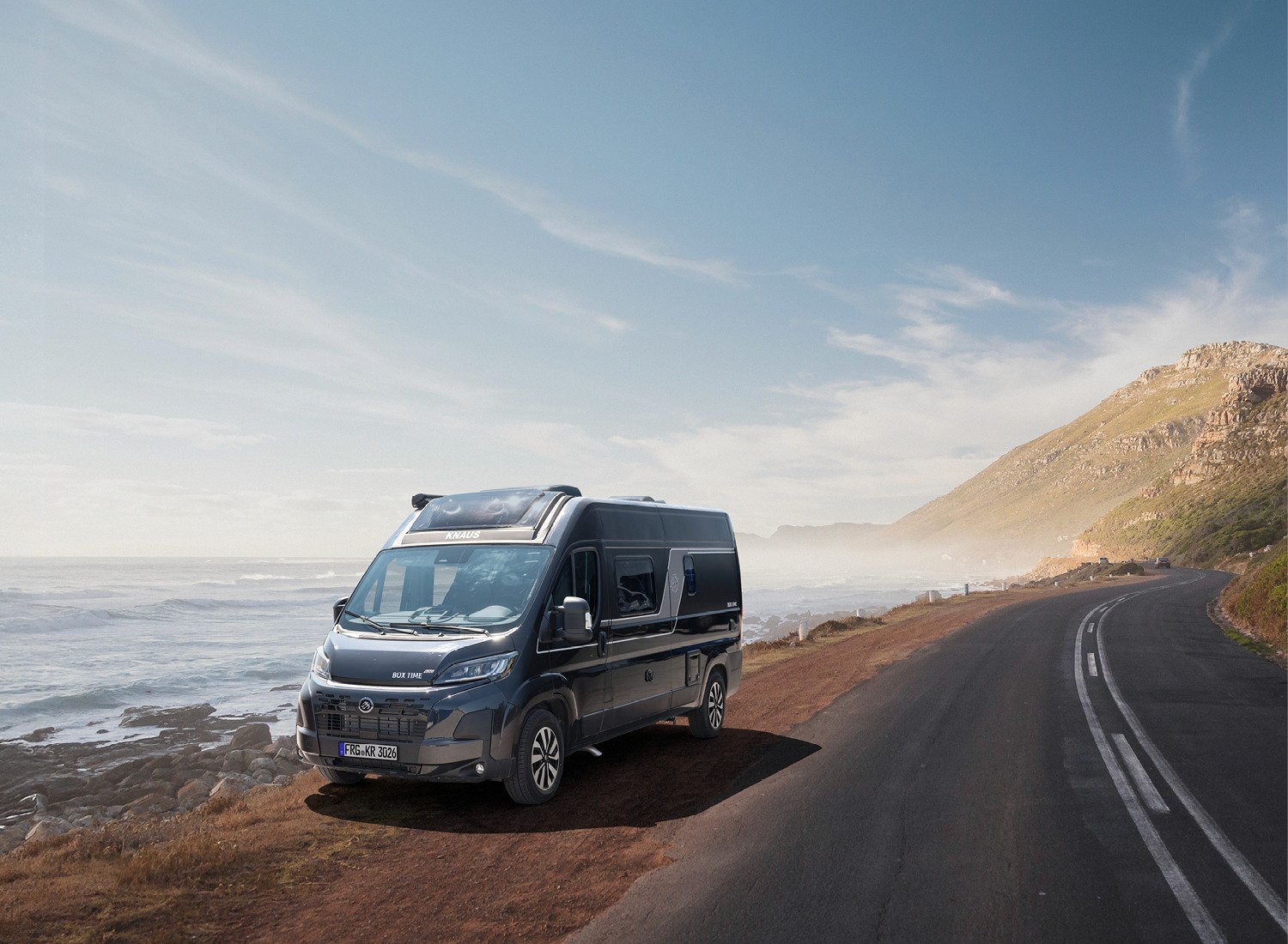 Ein KNAUS CamperVan steht an einer Küstenstraße mit Blick auf das Meer und umliegende Berge unter klarem Himmel.