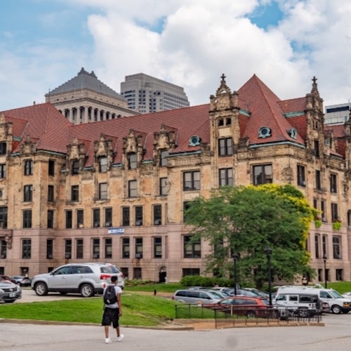 Historical building with red-tiled roofs, surrounded by parked cars and a few trees, with modern skyscrapers in the background.