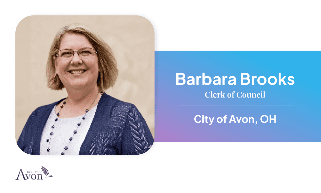 Female municipal clerk seated at office desk with nameplate reading 'Barbara Brooks, City of Avon, OH,' an American flag in the background, and office supplies and binders visible.