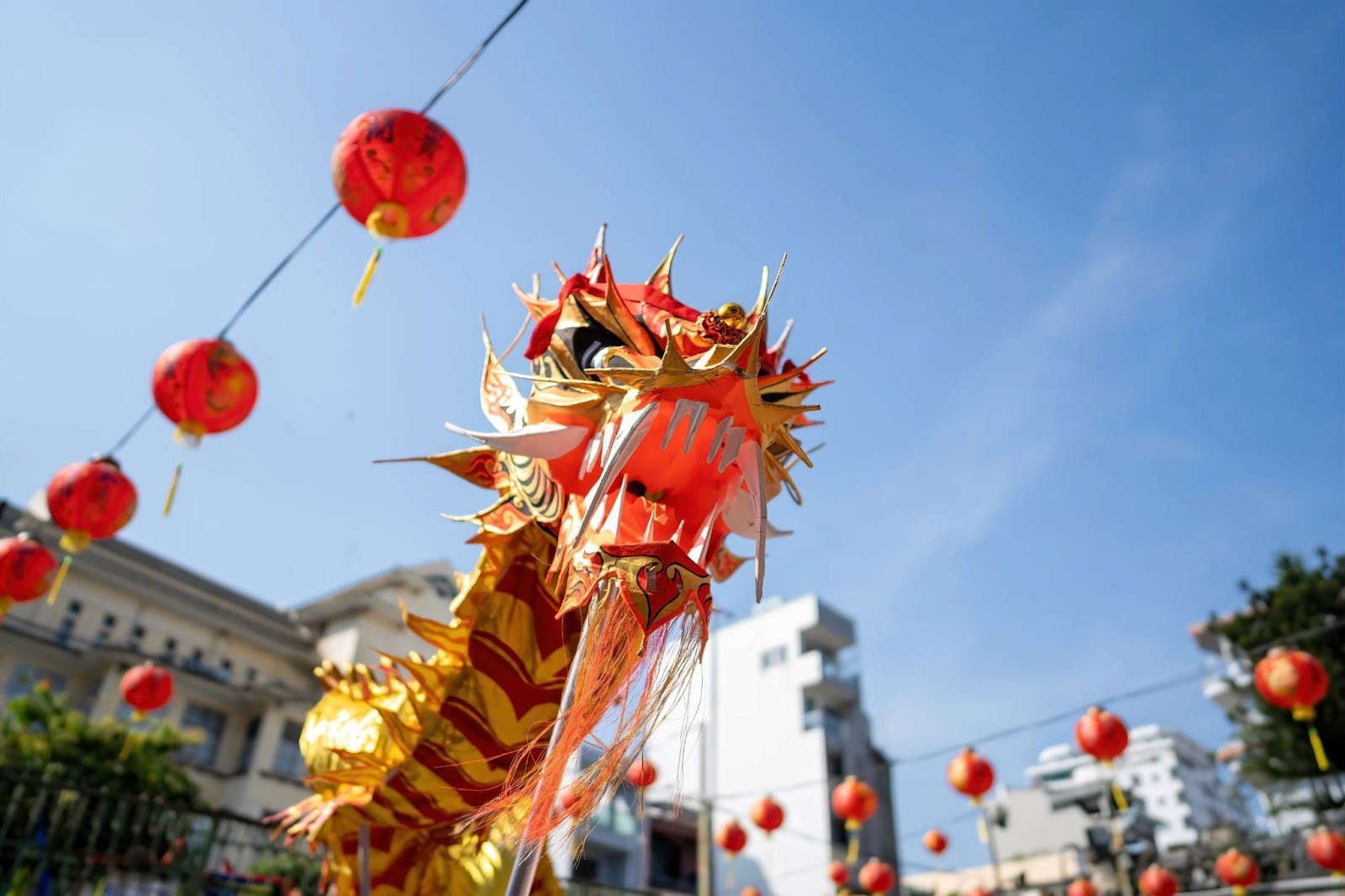 The lens is looking up at the head of a dragon puppet dancing in a square decorated with red lanterns, with blue sky beyond