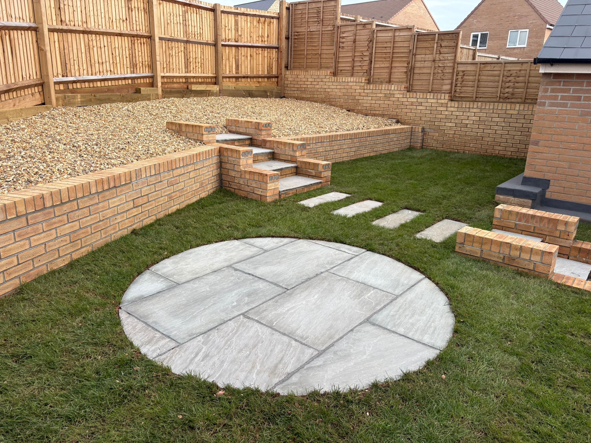 Circular paved seating area in a lawn, with brick retaining walls and steps leading to a gravel terrace.