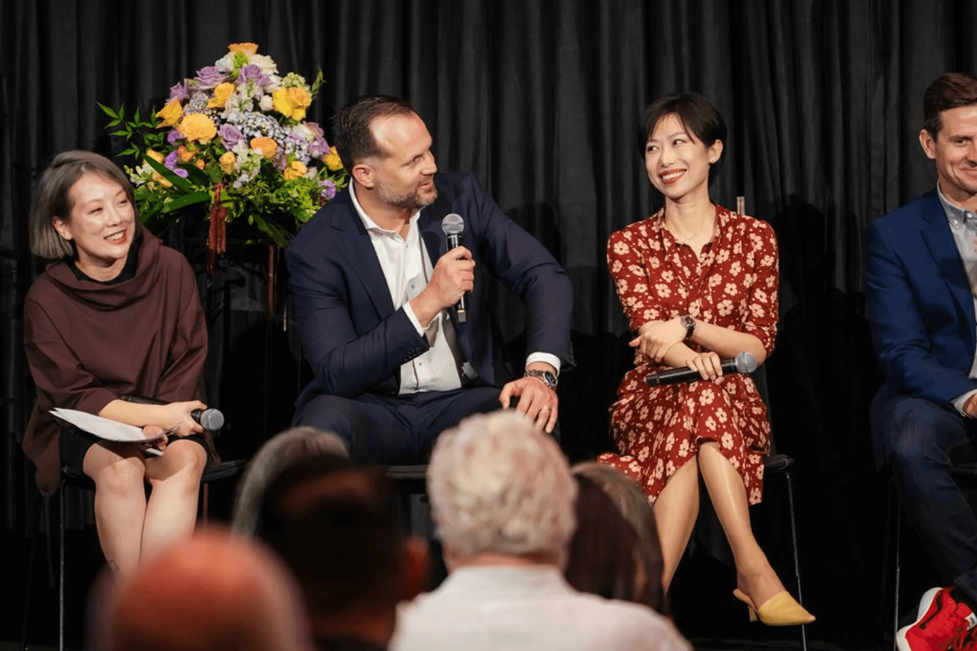 A panel of diverse speakers is seated on stage, engaging with an audience, featuring a smiling woman in a red floral dress, a man holding a microphone in a navy suit, and vibrant floral arrangements in the background, all set against a black curtain backdrop.