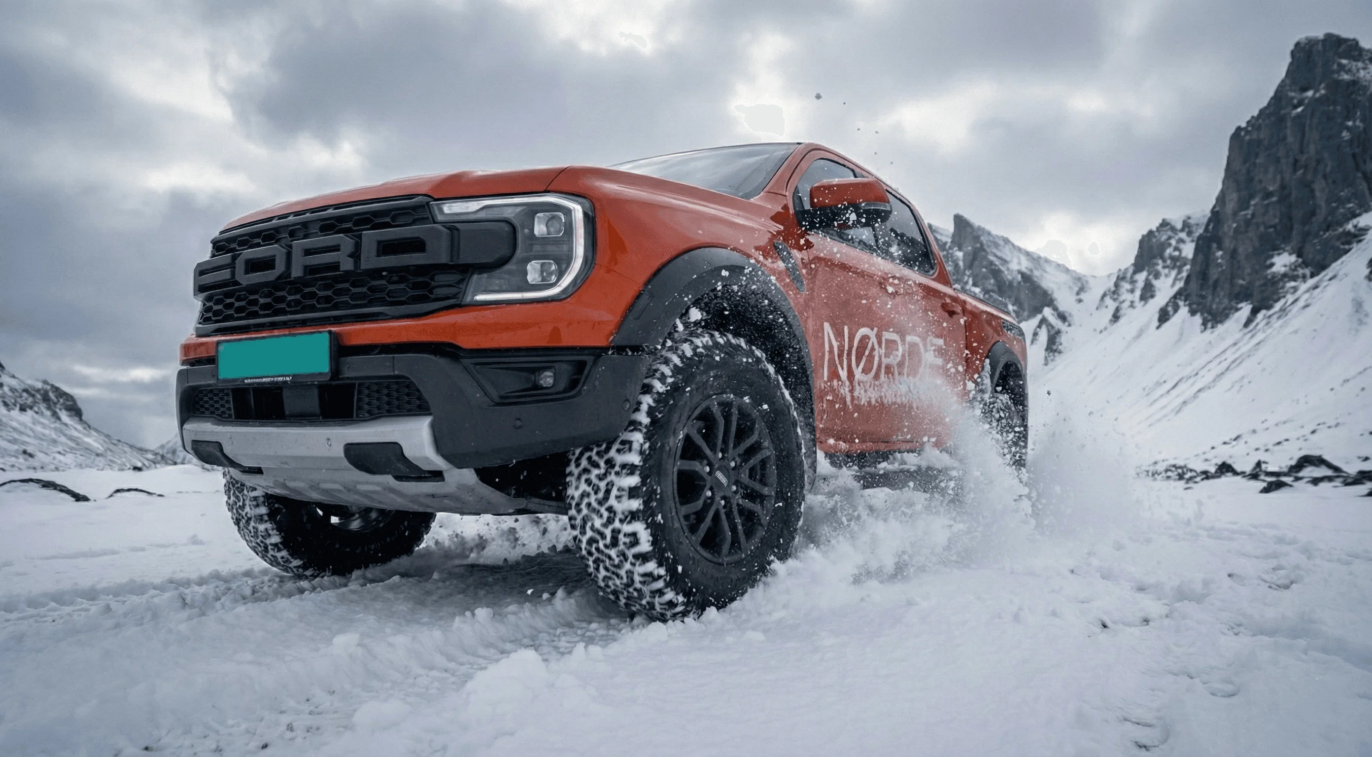 An orange Ford Ranger Raptor driving fast through deep snow, kicking up a cloud of white powder.
