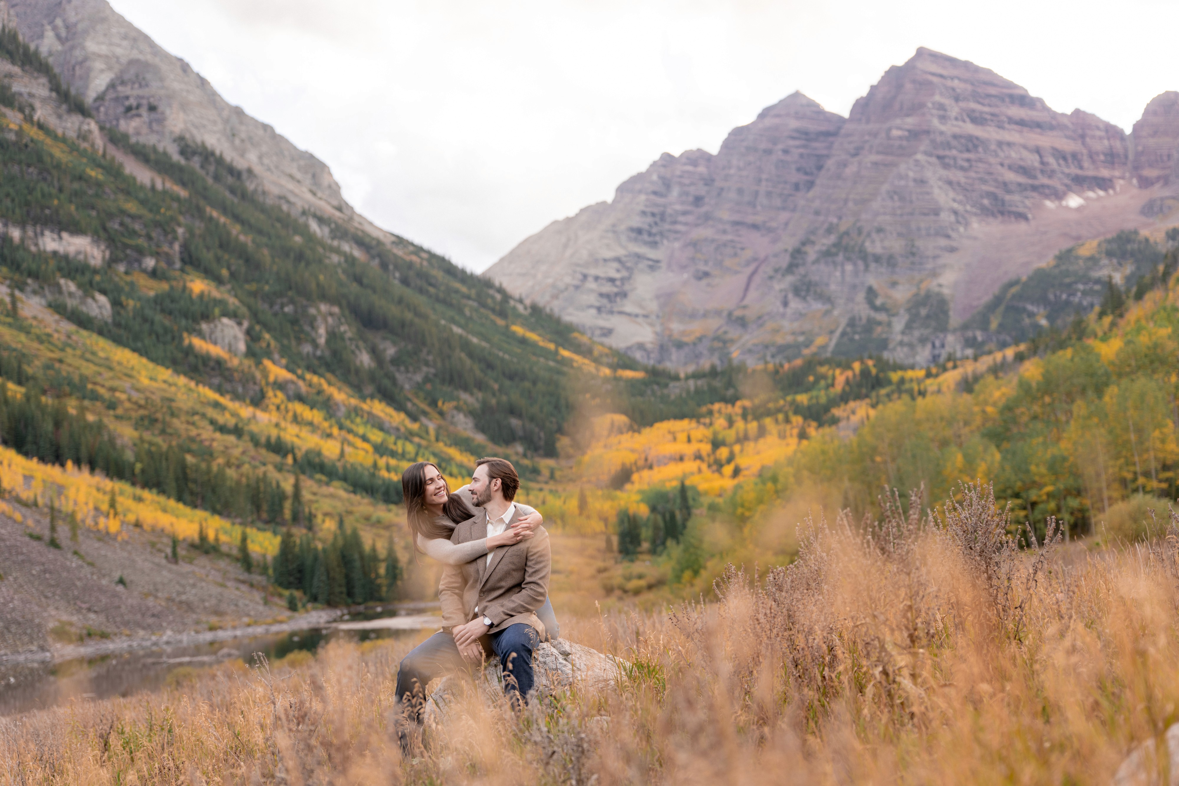 Tim and Lexi sit together on a rock in a meadow with Maroon Lake and the Maroon Bells behind them.