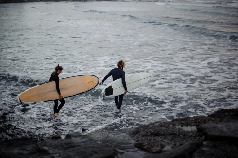 Two surfers in wetsuits carrying surfboard into ocean waves at coastal beach