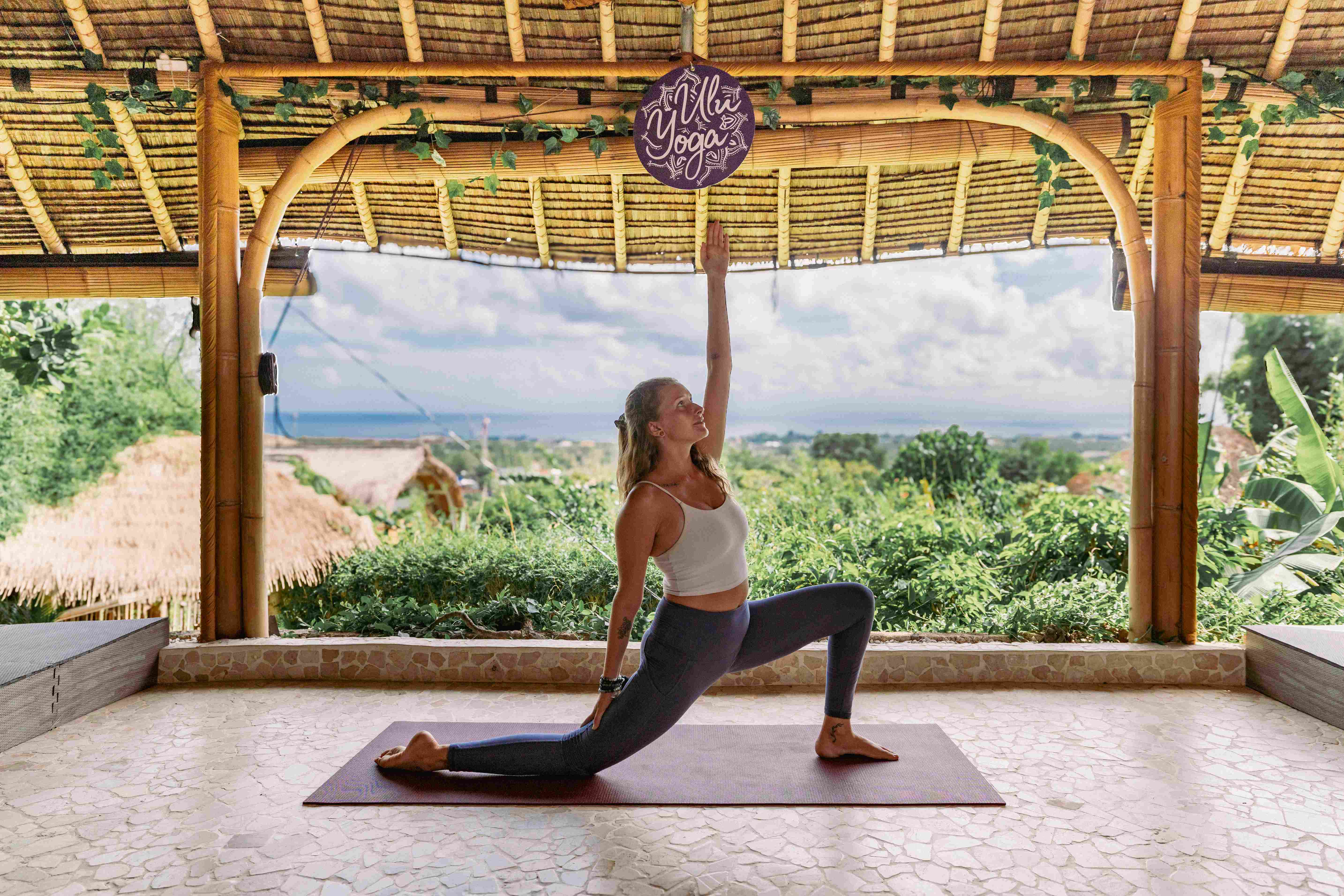 Yoga student practicing a High Lunge variation on the studio terrace with ocean view and Ulu Yoga sign.