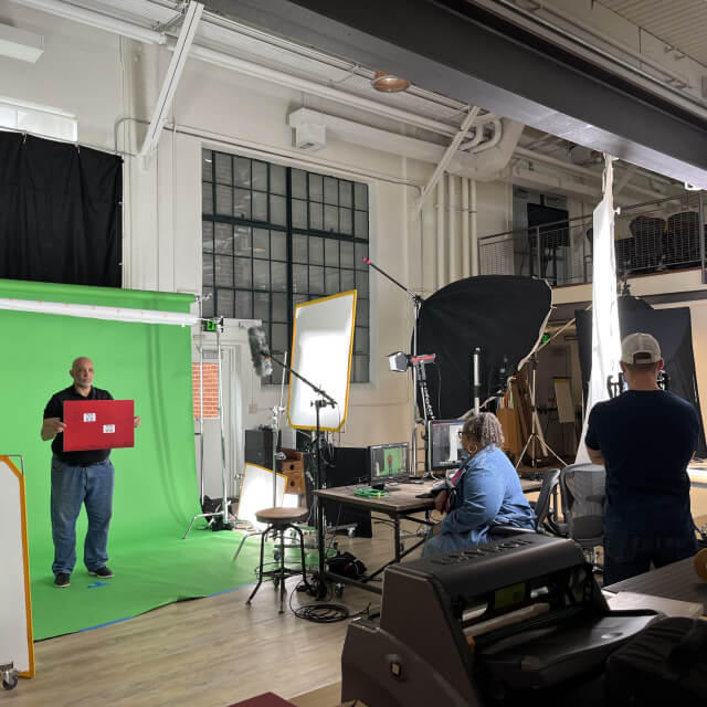 A photography studio setup with a green screen, lighting equipment, photographer at a computer, and a person holding a red sign in front of the green screen.
