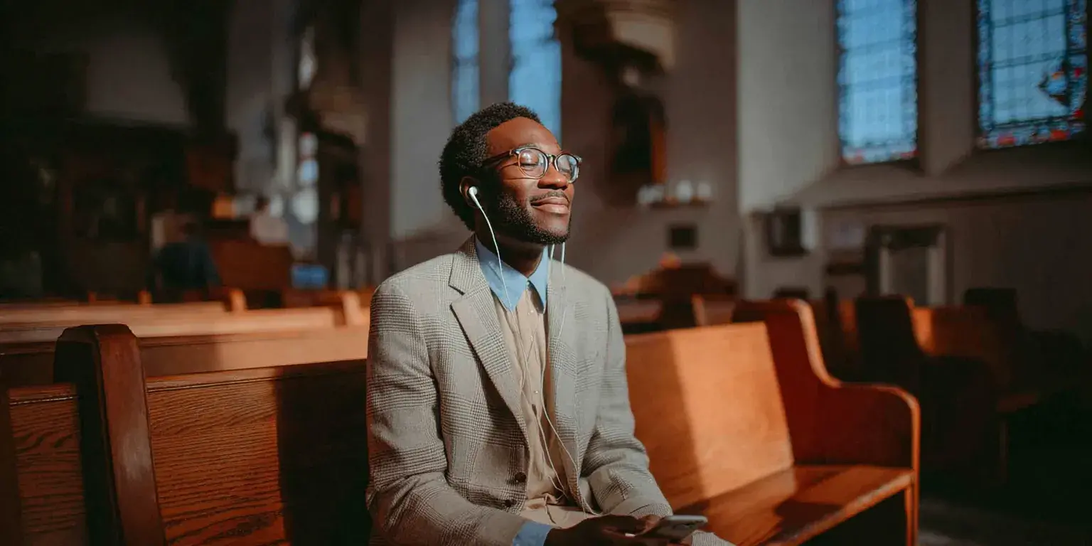 A man sitting in a church pew, smiling peacefully while listening to audio through earphones, symbolizing faith, reflection, and connection in a digital age.