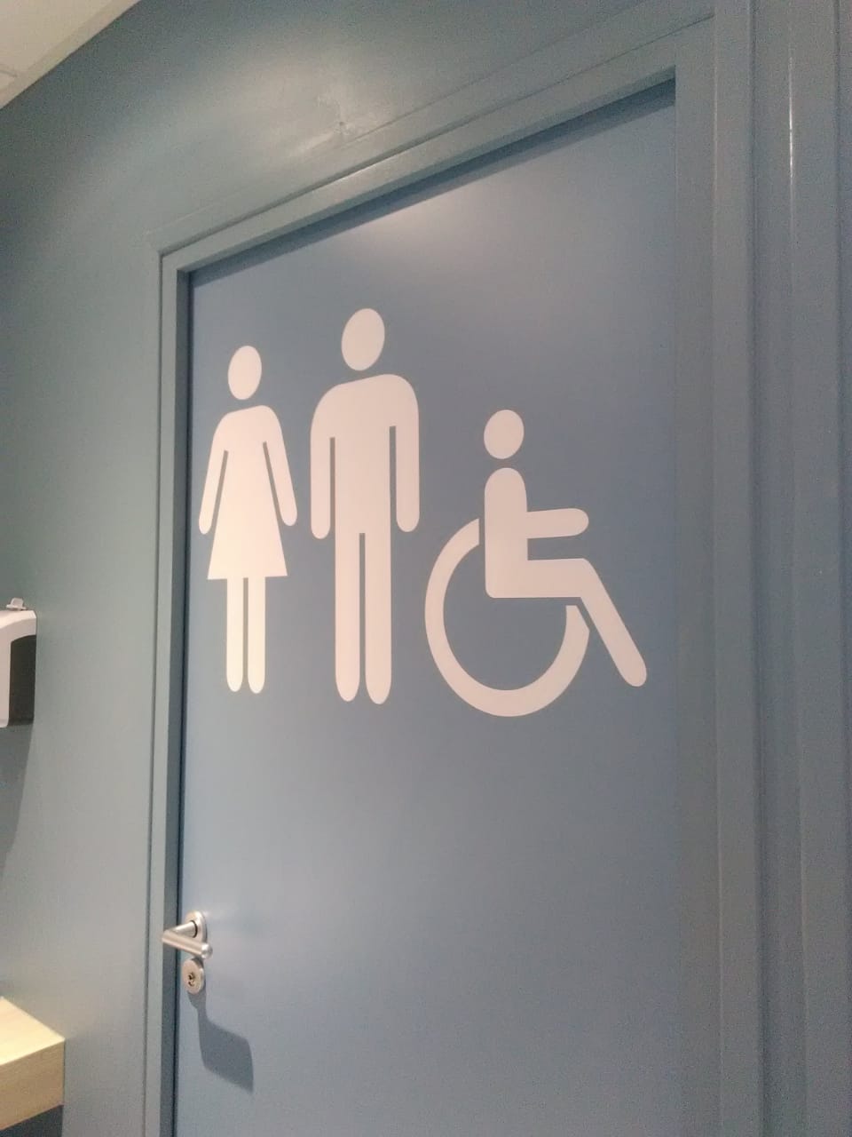 Porte bleue avec des icônes blanches représentant des femmes, des hommes et un fauteuil roulant, indiquant des toilettes mixtes et accessibles. Le cadre est propre et moderne.