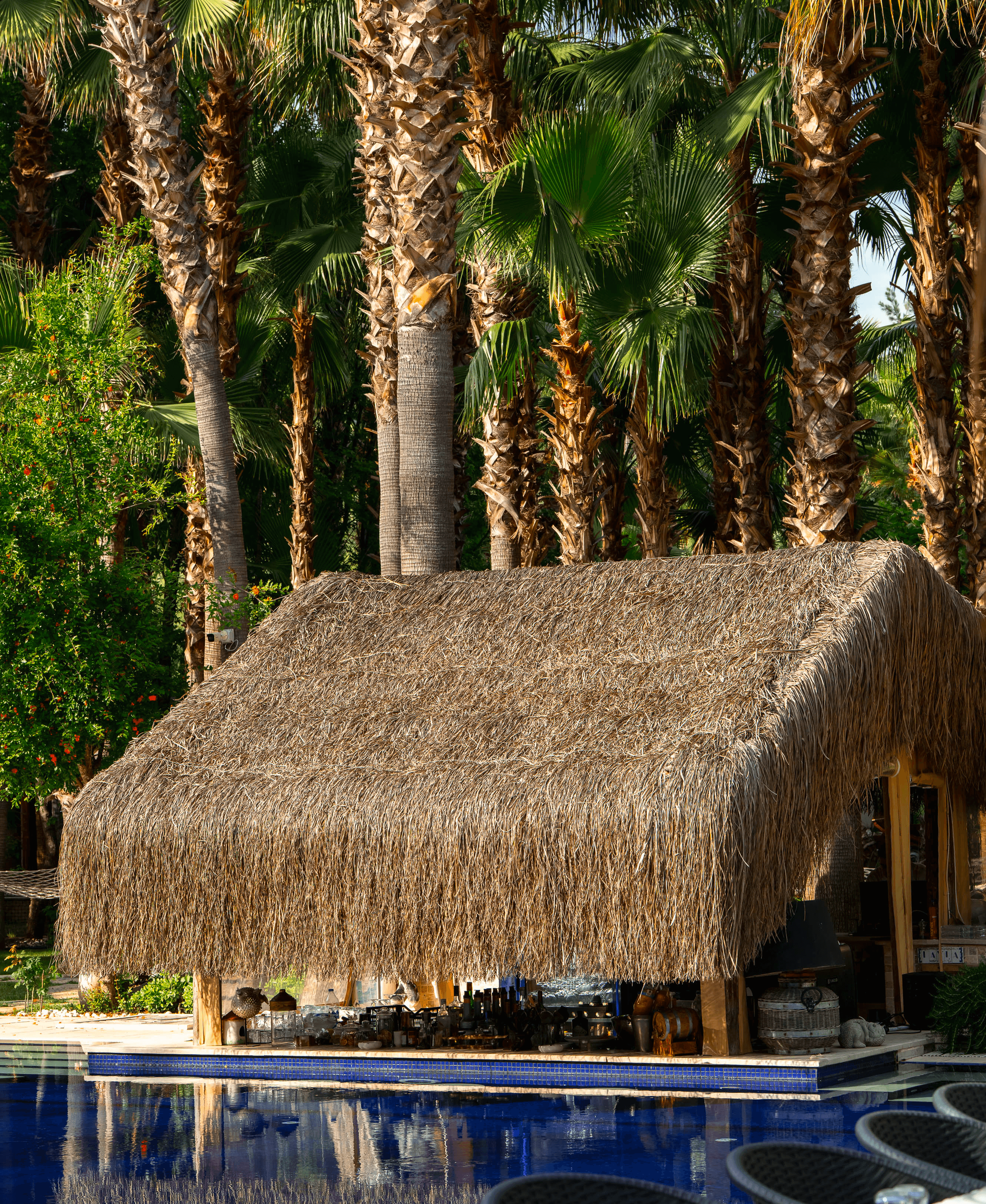 Outdoor lounge under a wooden pergola, draped linen curtains, beige cushions, ocean backdrop, palm shadows dancing on the walls, serene luxury.