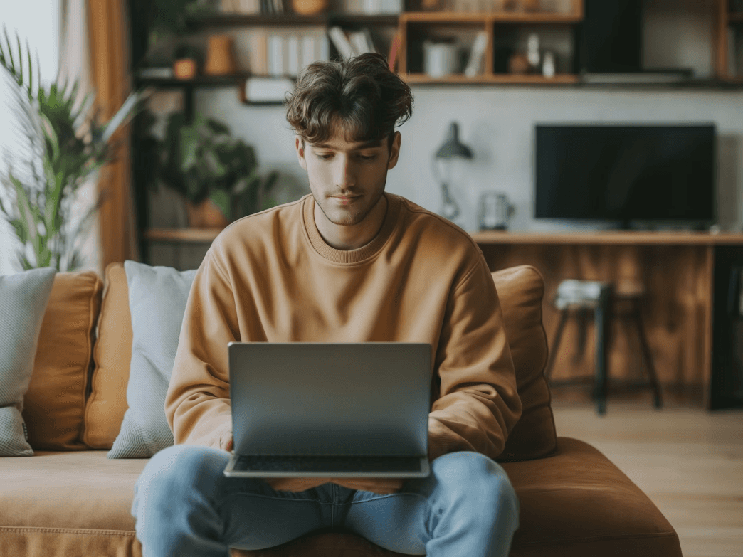 Young man in a tan sweater using a laptop on a couch.