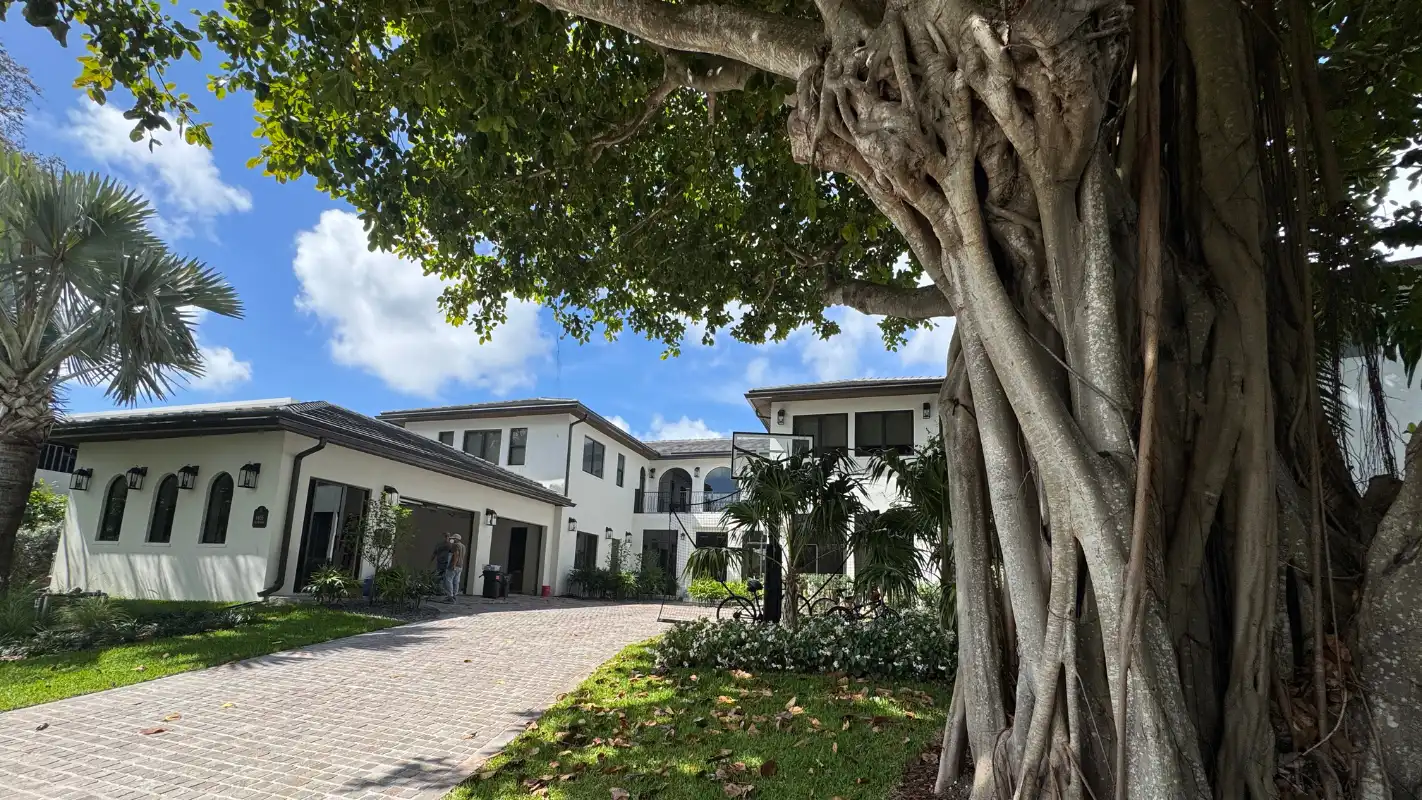 Front exterior of Island Road Residence on Bay Point Island, Miami — a Mediterranean Revival home with white stucco facade, arched windows, tile roof, and brick driveway framed by a mature banyan tree, by Atelier International.