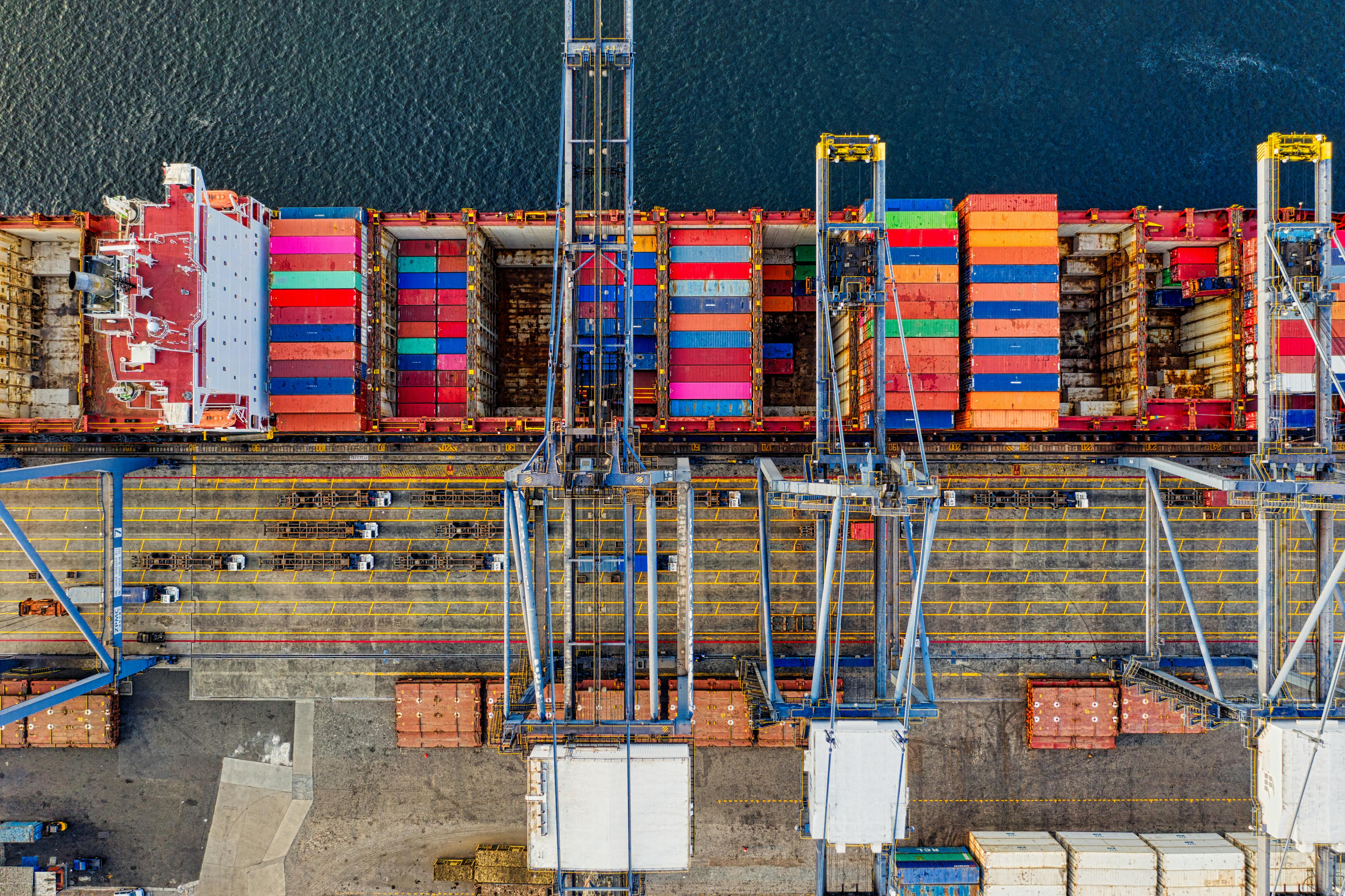 Aerial view of a large container ship docked at a port, filled with brightly colored shipping containers, with cranes and trucks visible on the quay.