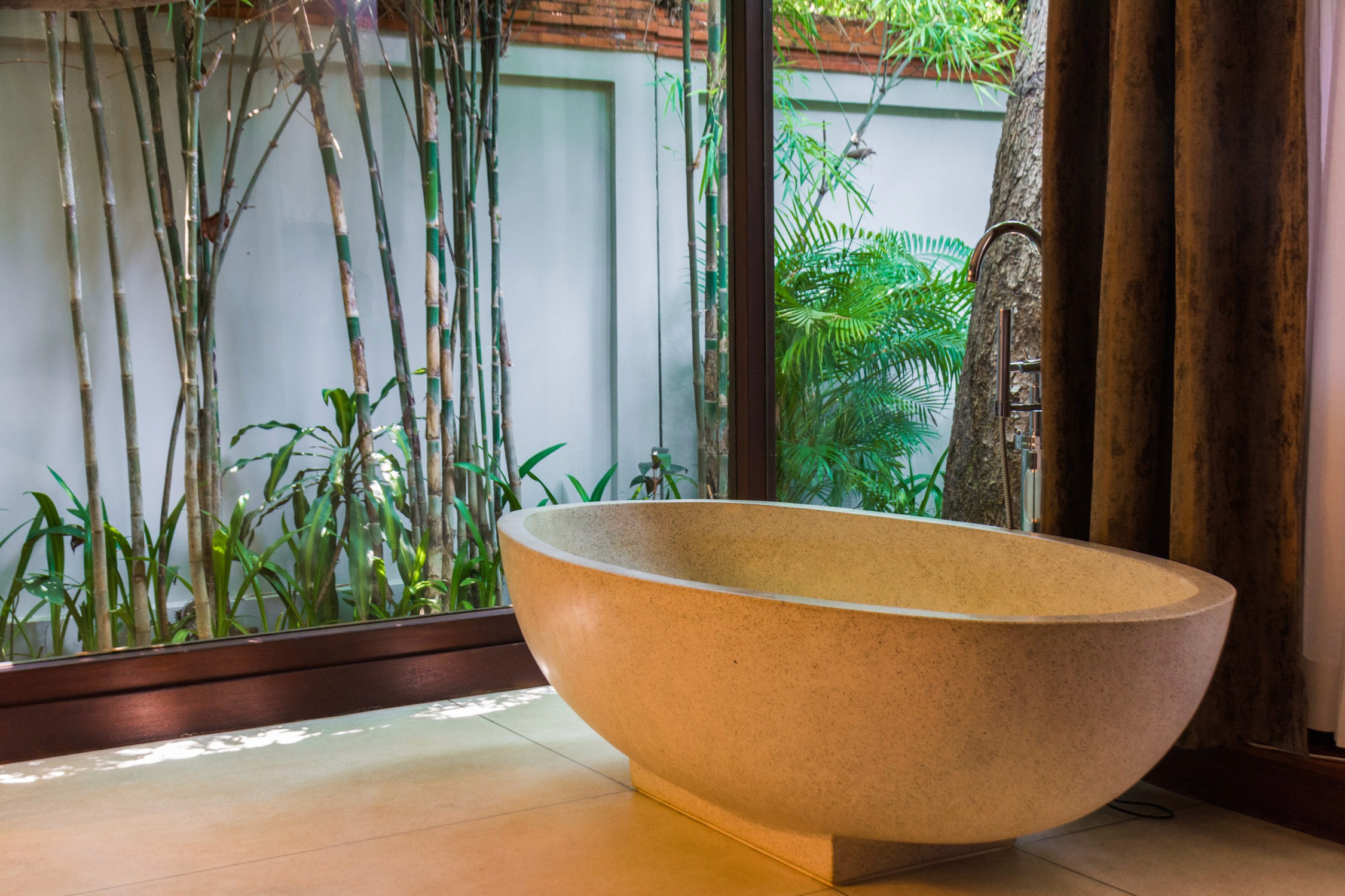 A stone soaking tub sits in a spa room with bamboo stalks and tropical greenery visible through a glass wall.