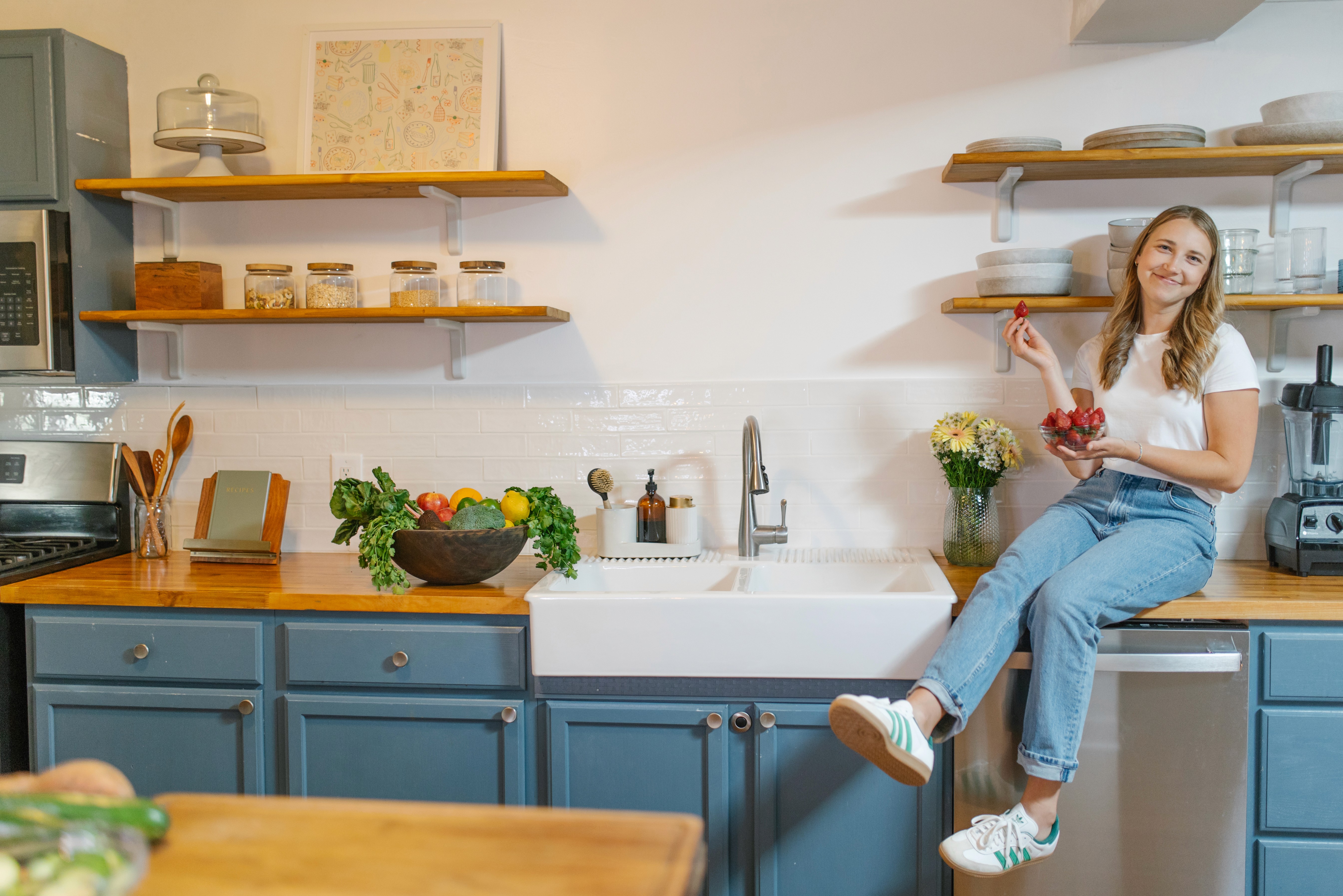 A smiling woman sits atop a wooden countertop in a kitchen with a white tile backsplahs and organized glass jars and dishware on shelves