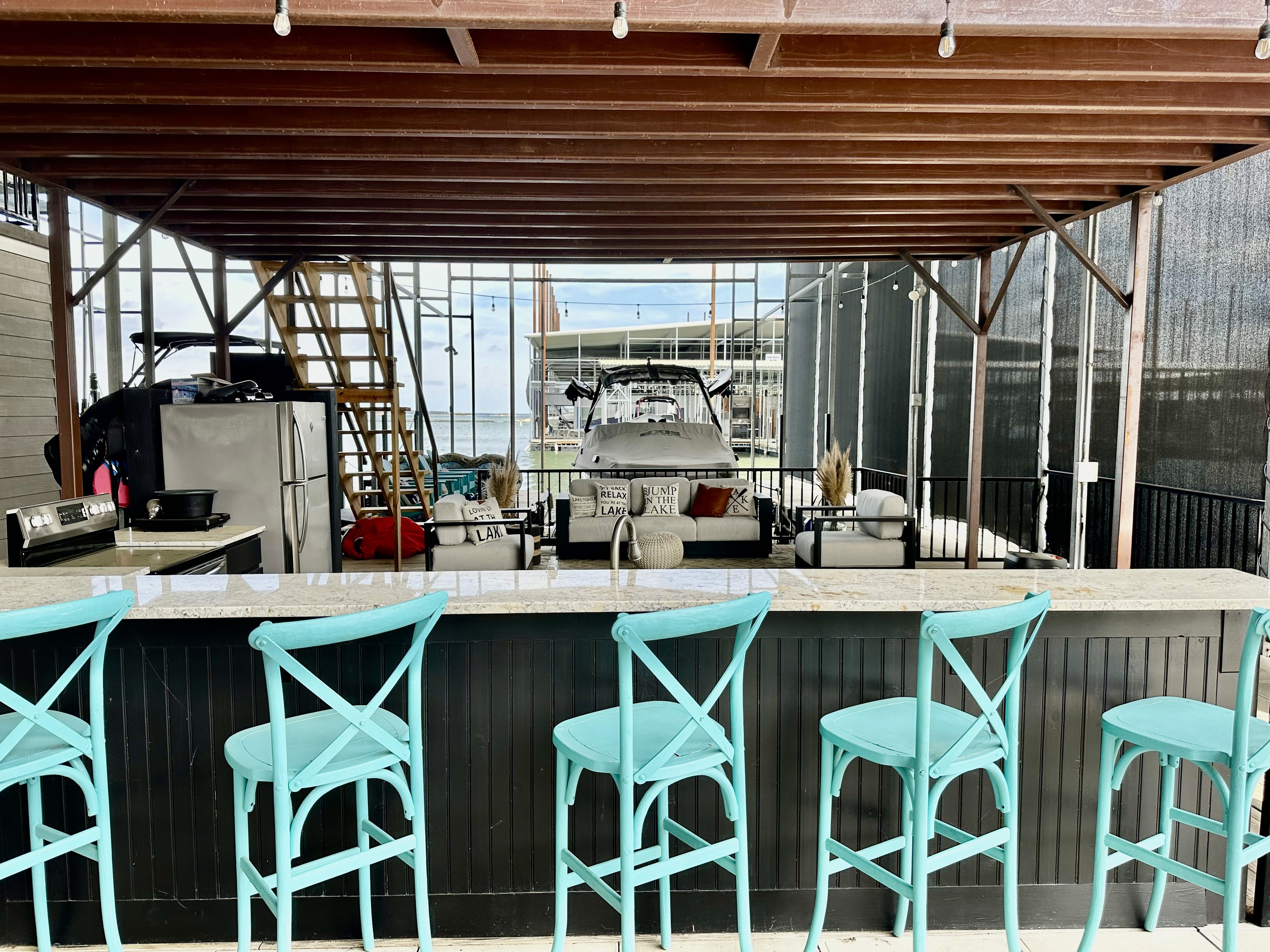 Outdoor bar area with turquoise bar stools, a stone countertop, and a covered seating area overlooking a marina with boats in the background.