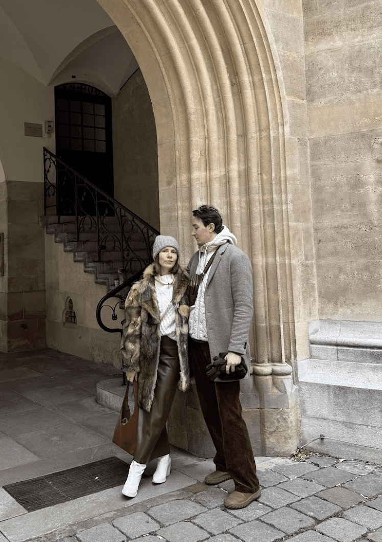 A couple stands near a stone archway, engaged in conversation, dressed in stylish outfits.