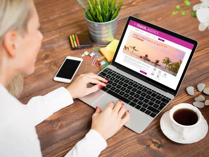 A person is seen from above, browsing a travel agency website on their laptop, which displays an inviting tropical resort offer. The setting includes a neatly organized desk with a smartphone, colorful stationery, a potted plant, and a cup of coffee, indicating a comfortable and well-arranged workspace.