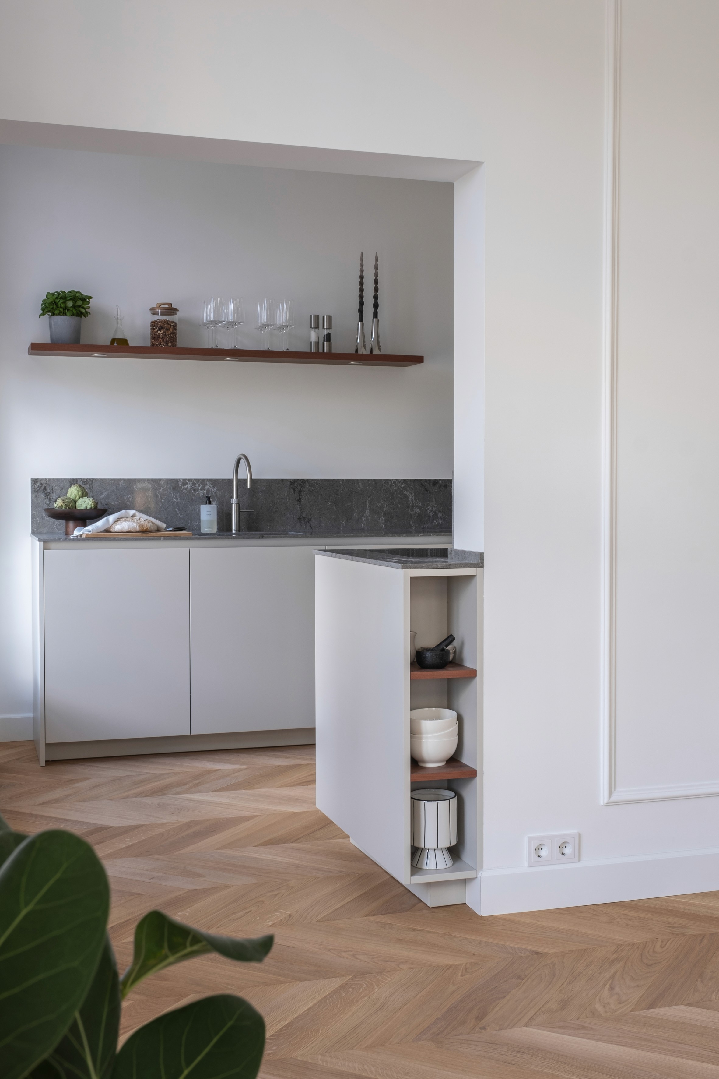 Minimalist kitchen niche with grey stone backsplash, walnut floating shelf with glassware, light grey handleless cabinetry, and Hungarian Parquet parquet floor