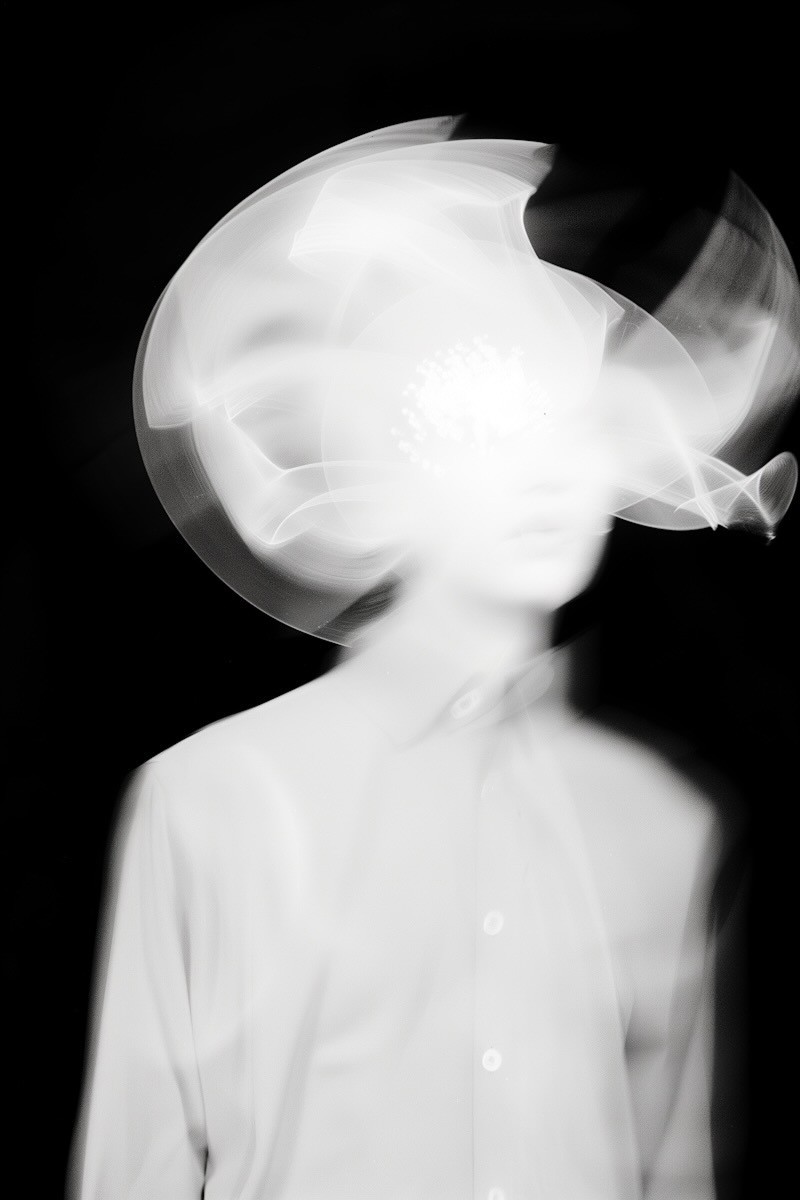 Abstract black-and-white image of a blurred, ghostlike figure in a white shirt, with a glowing, smoky head against a black background.