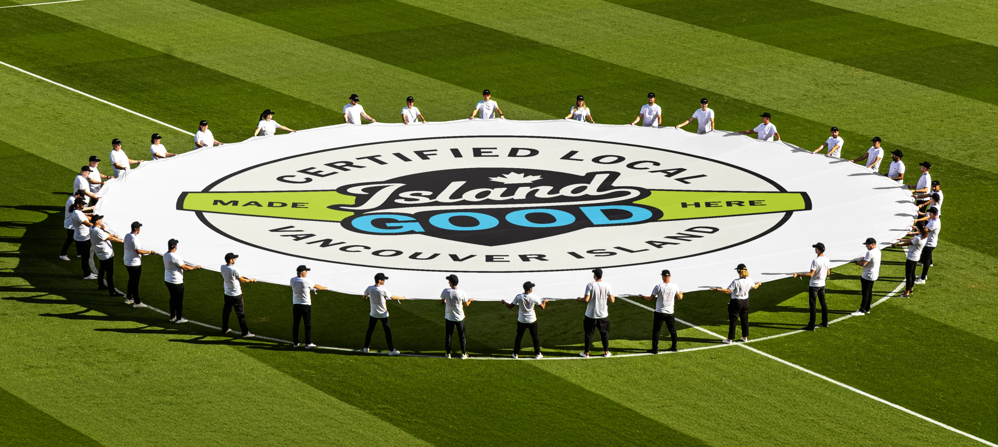 A large circular banner on a sports field being held by a group of people. The banner has an Island Good badge printed large on the top that says "Certified Local Vancouver Island"