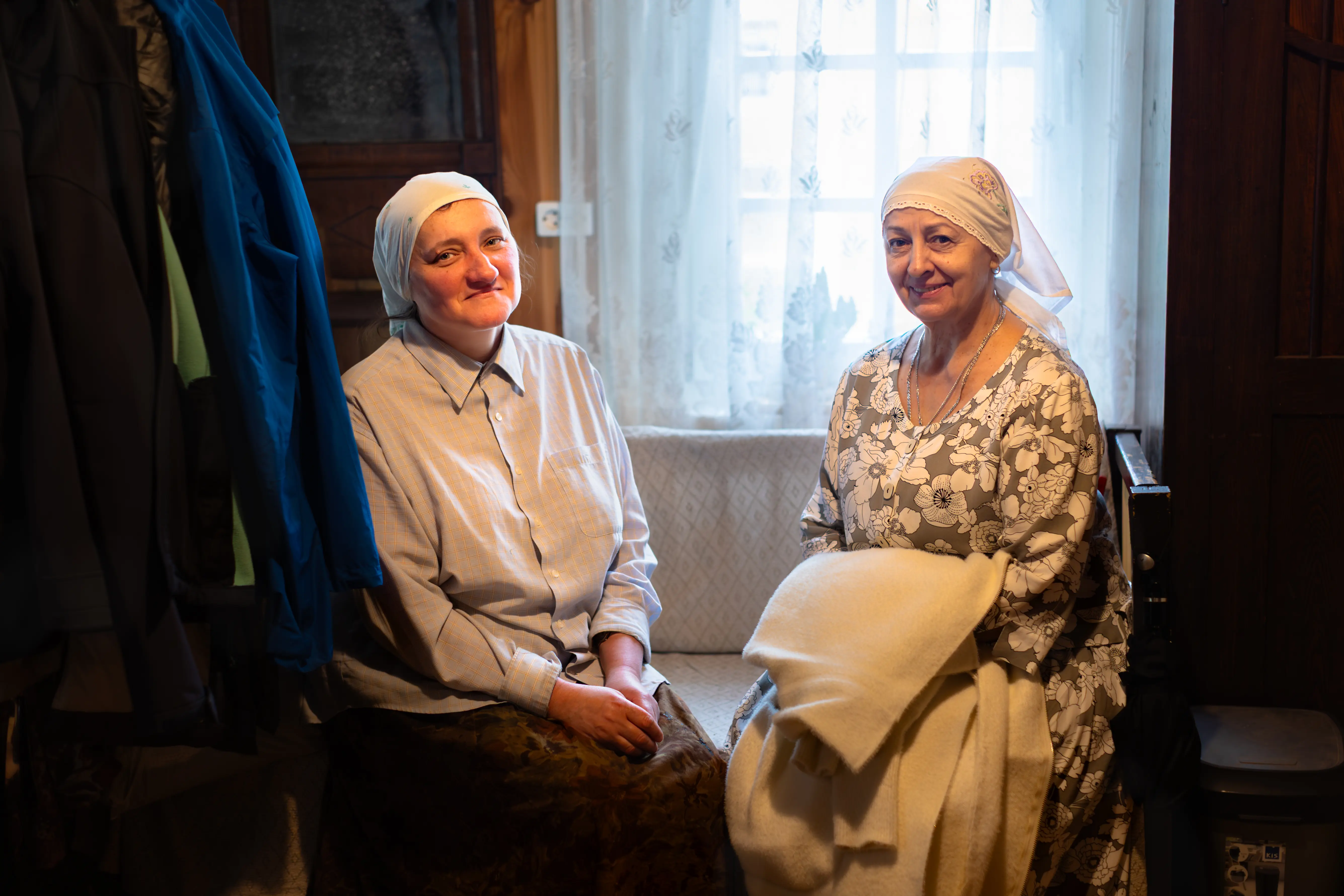 Dwie kobiety siedzące na ławce w jasnym wnętrzu, obie ubrane w nakrycia głowy. Jedna z nich ma na sobie szary strój, a druga suknię w kwiaty. W tle widać okno z firanką.