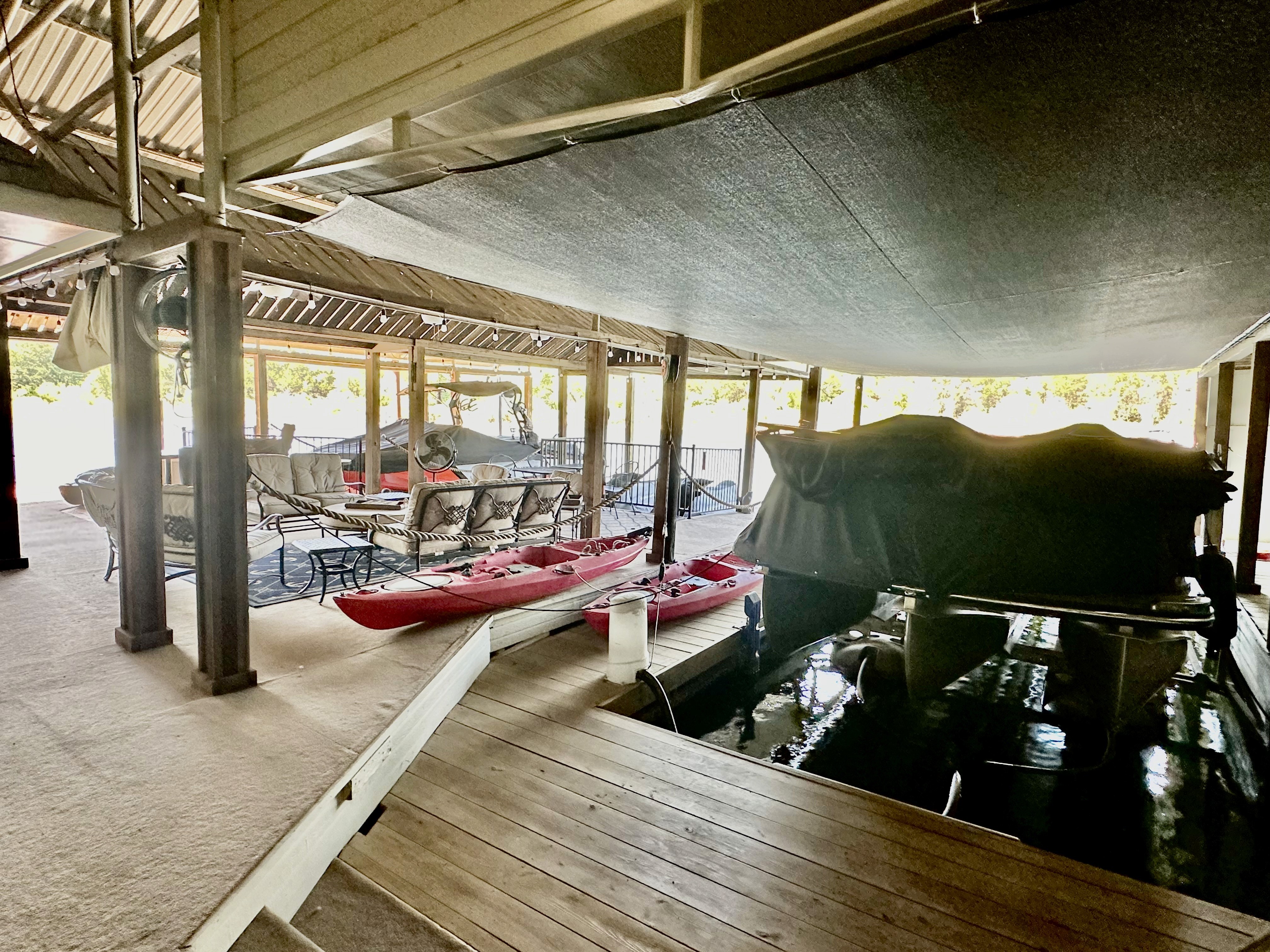 A spacious, covered outdoor area features patio furniture, two red kayaks on a wooden deck, and a small boat docked in an adjacent slip, with ceiling shade and natural light filtering through.