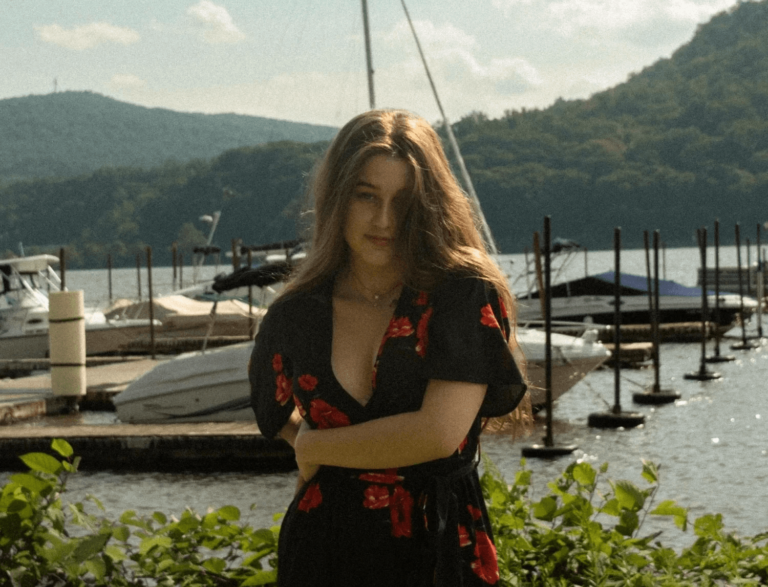 Varvara Aristakesyan stands at a marina wearing a black floral dress with red roses, arms crossed, looking directly at the camera with a calm expression. Sailboats, a dock, and green hills are visible in the background.