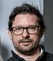 A man with dark hair and a beard, wearing black-rimmed glasses, looks directly at the camera embodying the expertise.