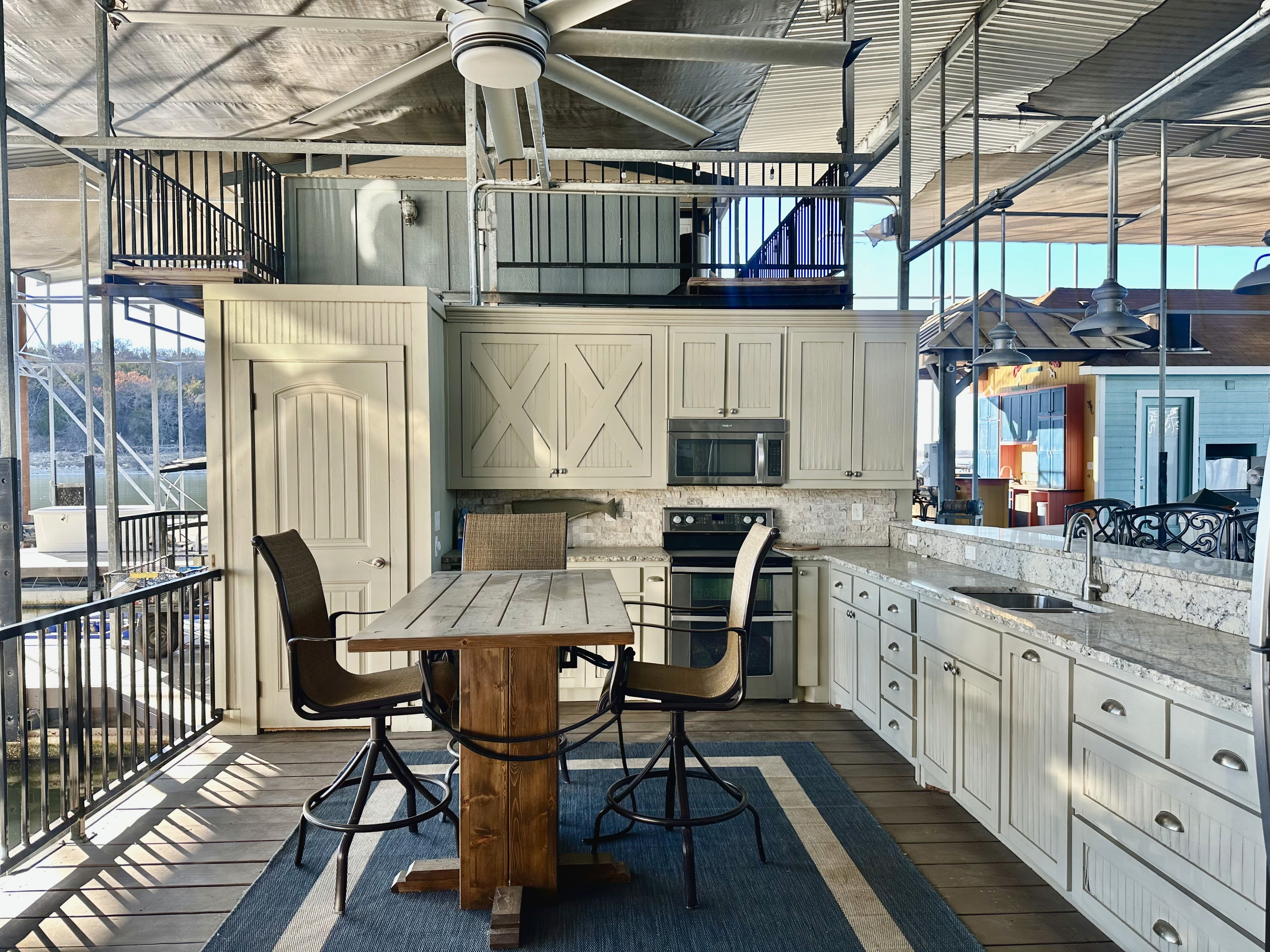 A modern outdoor kitchen on a lakeside deck features white cabinetry, a large wooden dining table with wicker barstools, stainless steel appliances, and an overhead ceiling fan under a metal canopy, all with a view of the lake in the background.