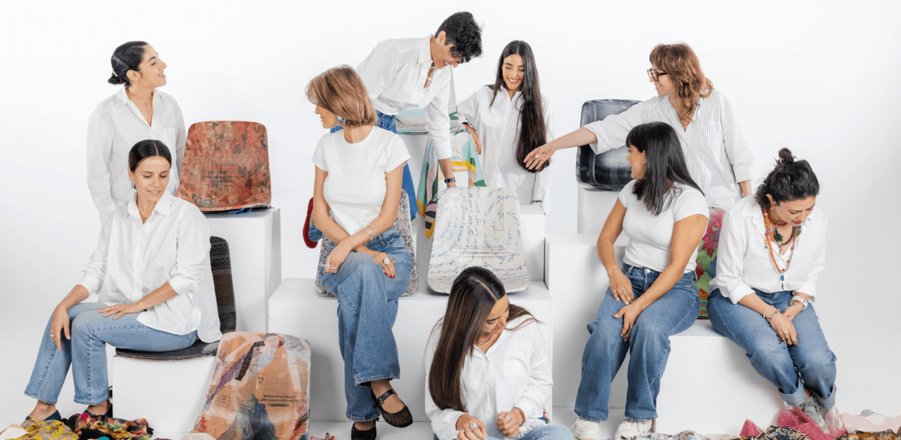 Group portrait of Iranian and non-Iranian women involved in Crafted Liberation, seated together on plinths surrounded by donated headscarves and fabric-covered stools.