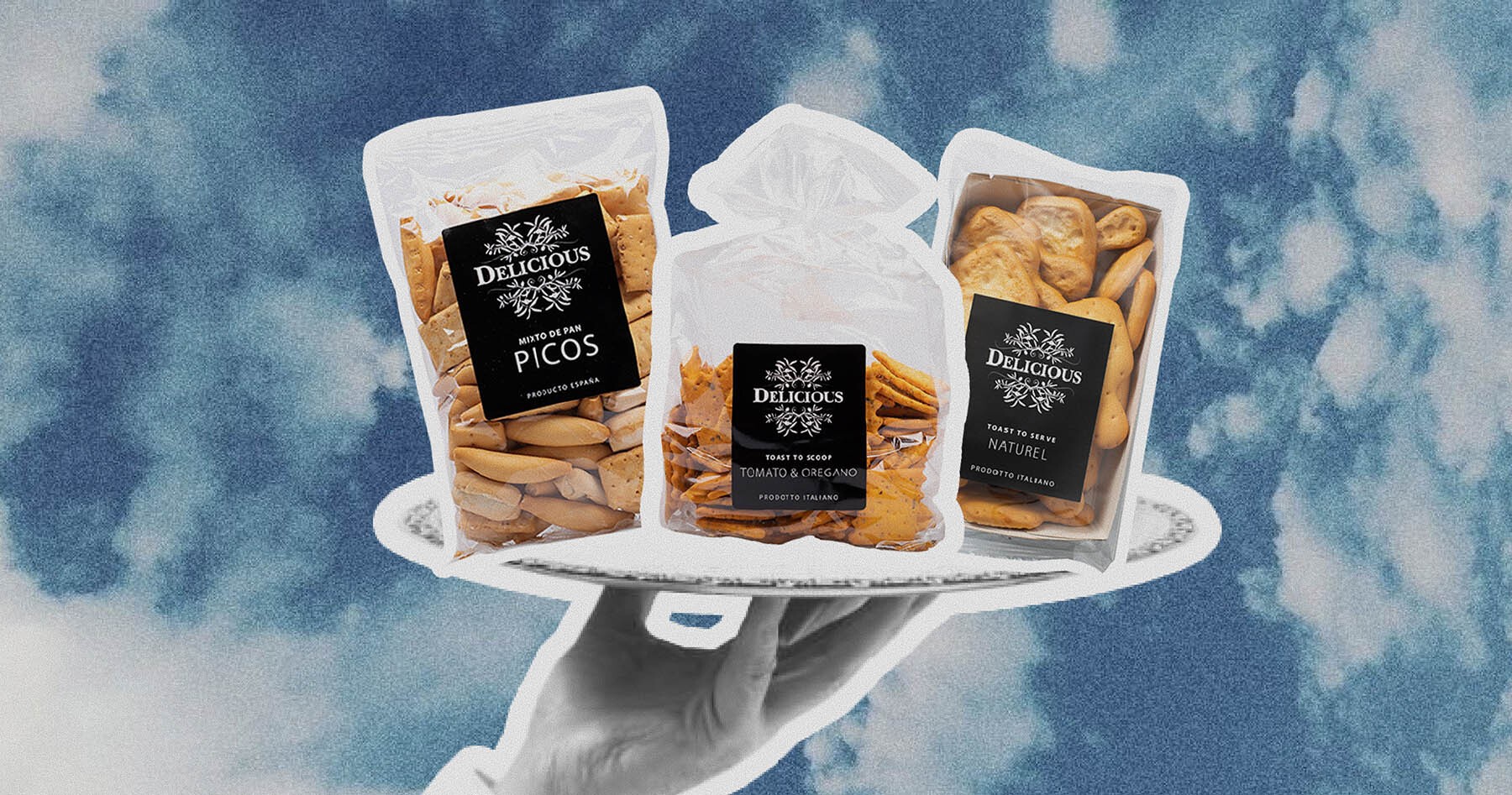 A hand holding a tray with three packages of "Delicious" branded Spanish and Italian snacks (Picos, Tomato & Oregano crackers, Naturel), perfect for gourmet gift boxes.
