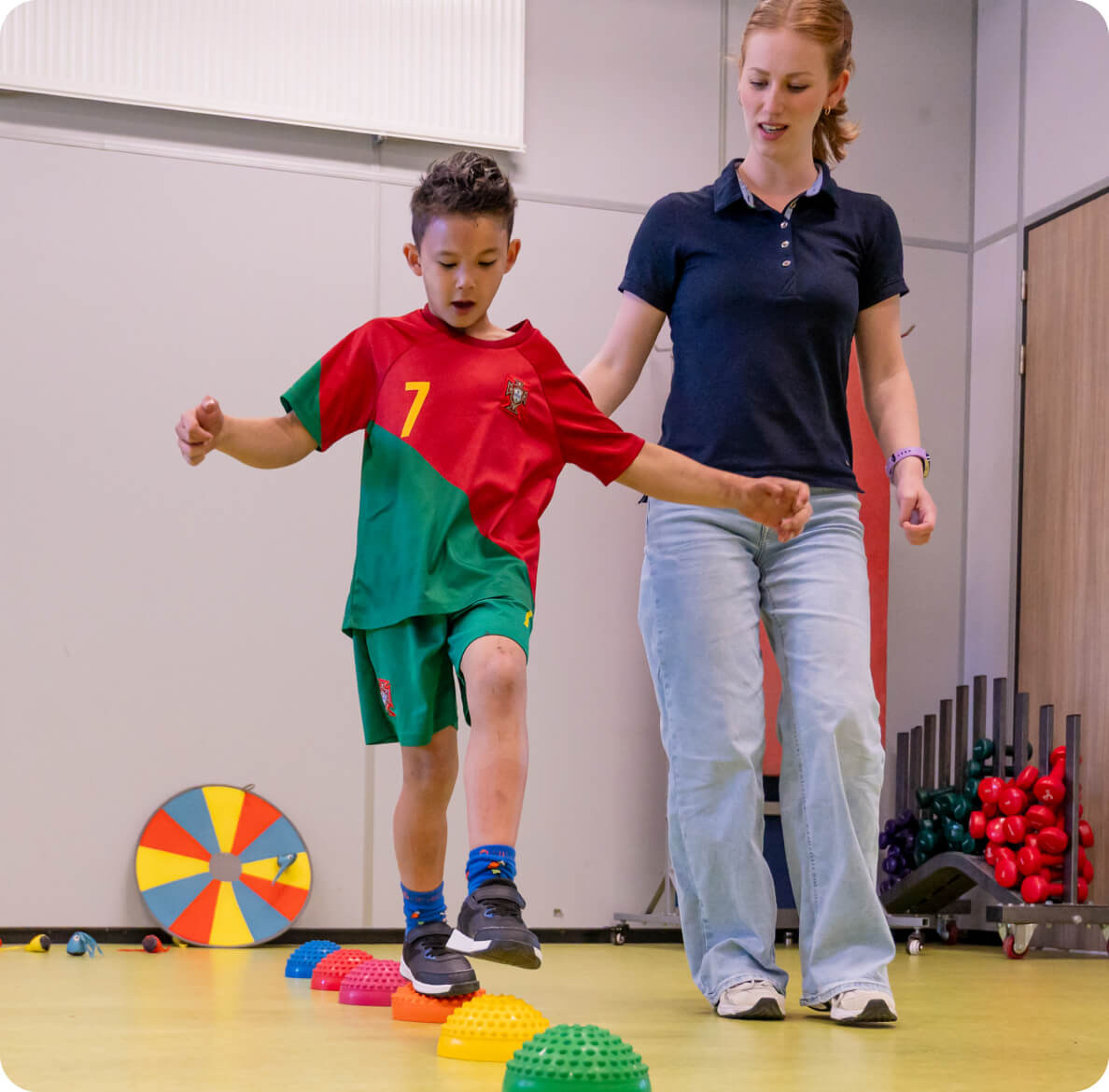A young child in a green and red sports outfit carefully balances on colorful stepping stones, guided by a woman in a navy shirt, on a brightly lit indoor play area.