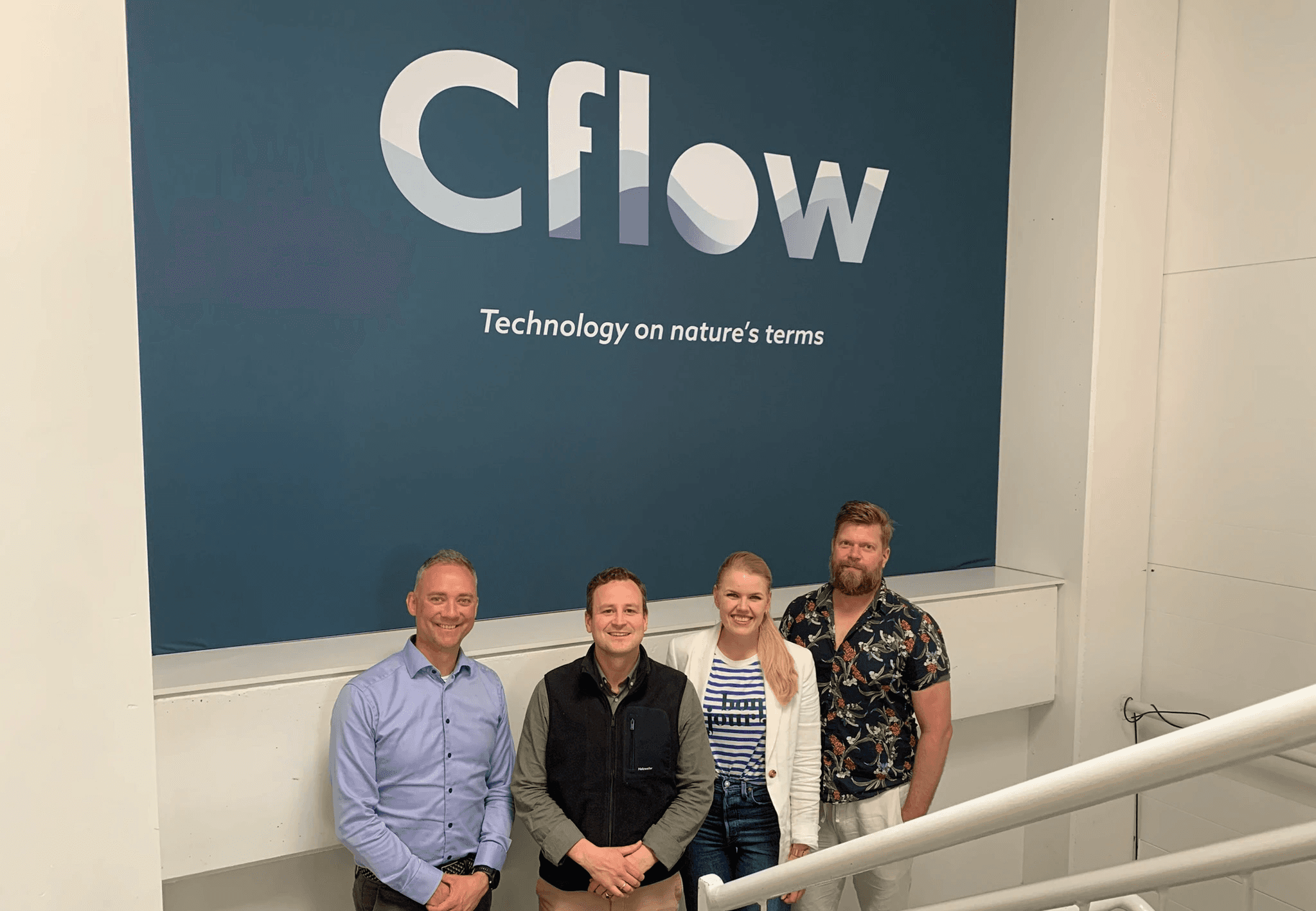 Group of four people posing in front of a blue wall with the Cflow logo in a modern office setting.